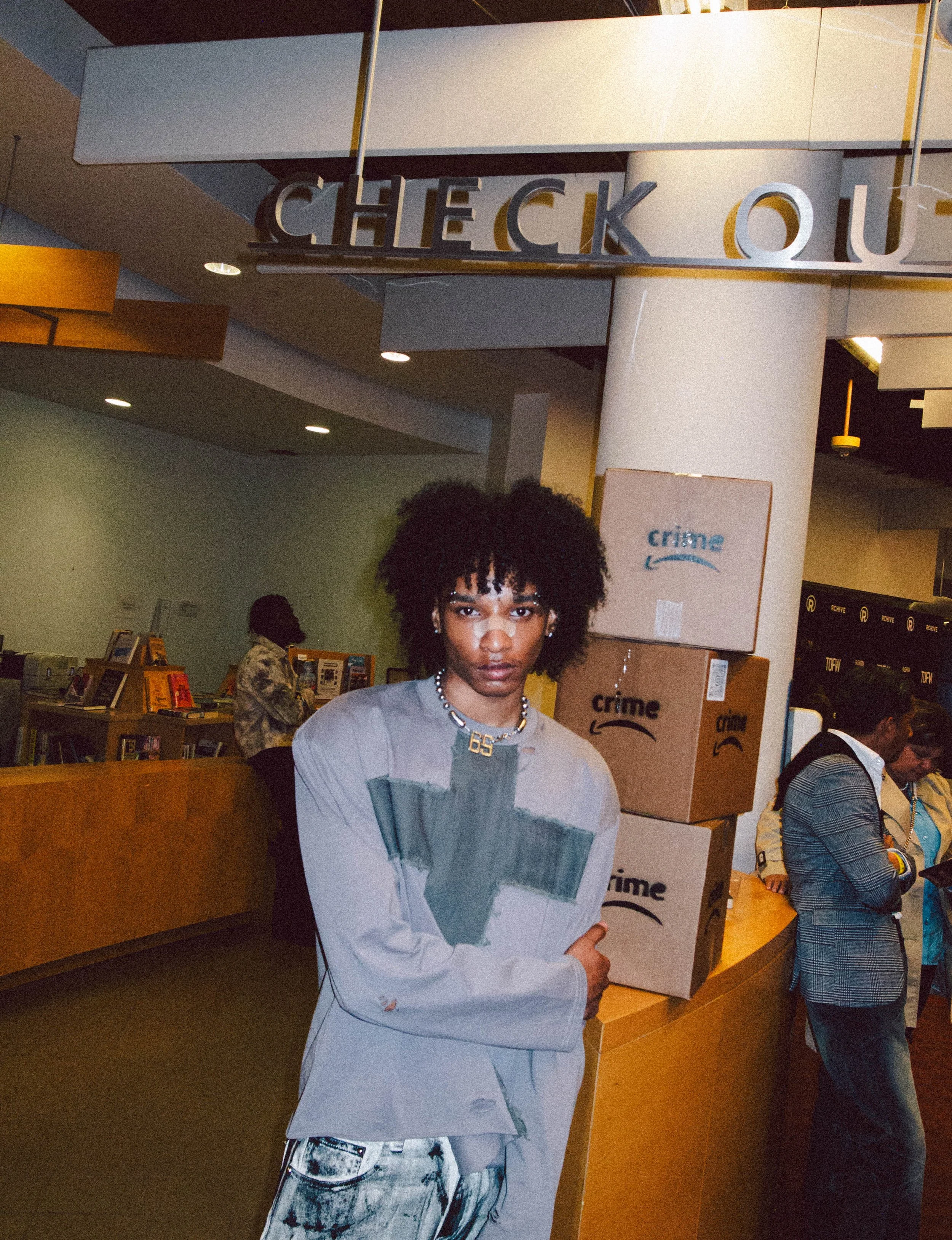 A young woman with curly hair standing at an Amazon bookstore check-out area, with cardboard boxes marked "crime" behind her, and other people shopping in the background.