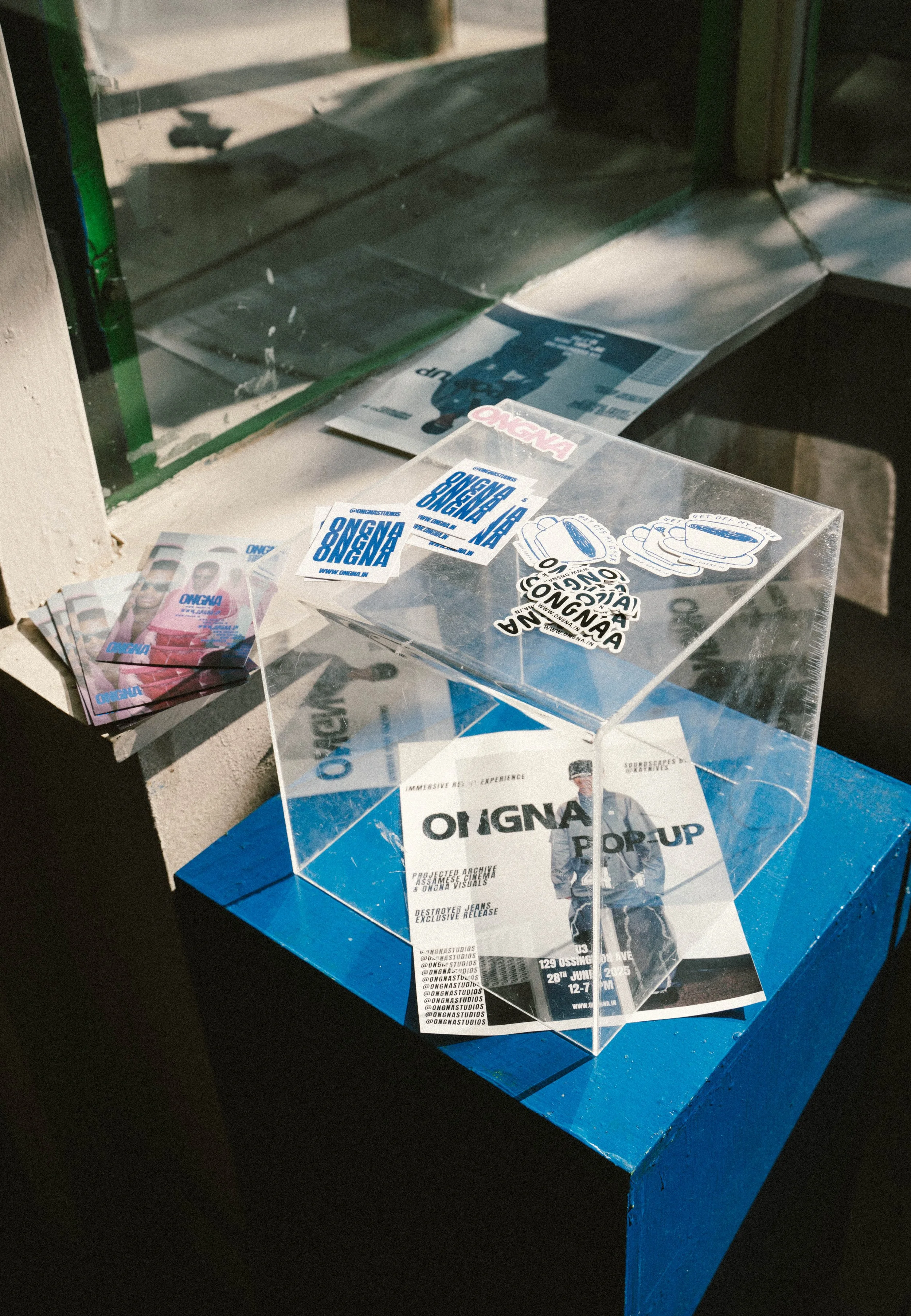 Flyers and stickers for an event called 'Ongna Pop Up' inside a transparent display case and on a blue table near a window.