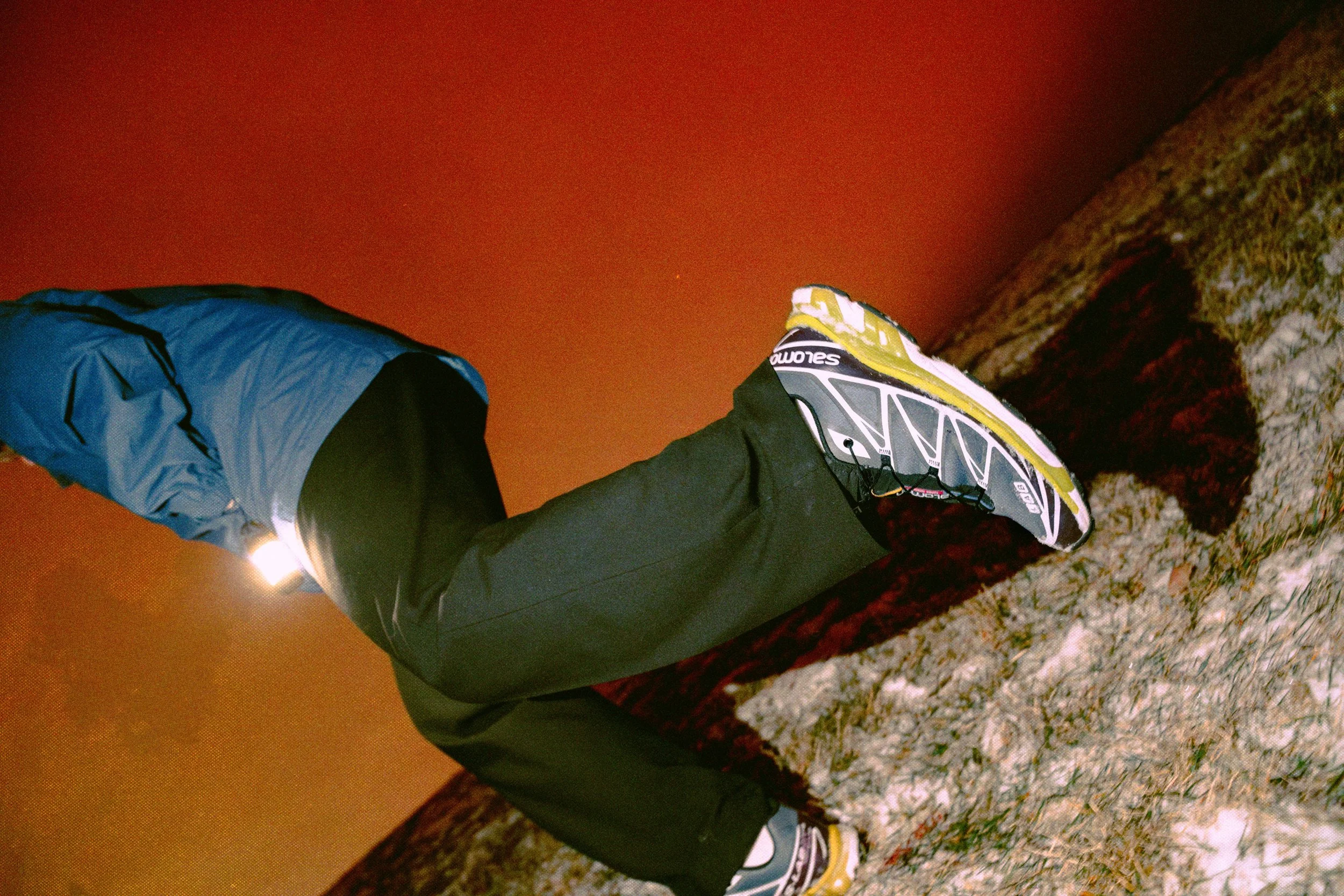 A person in outdoor gear, including a headlamp, and wearing a pair of Salomon xt6 is leaning over a snow-covered rocky surface at night, with an orange sky in the background.