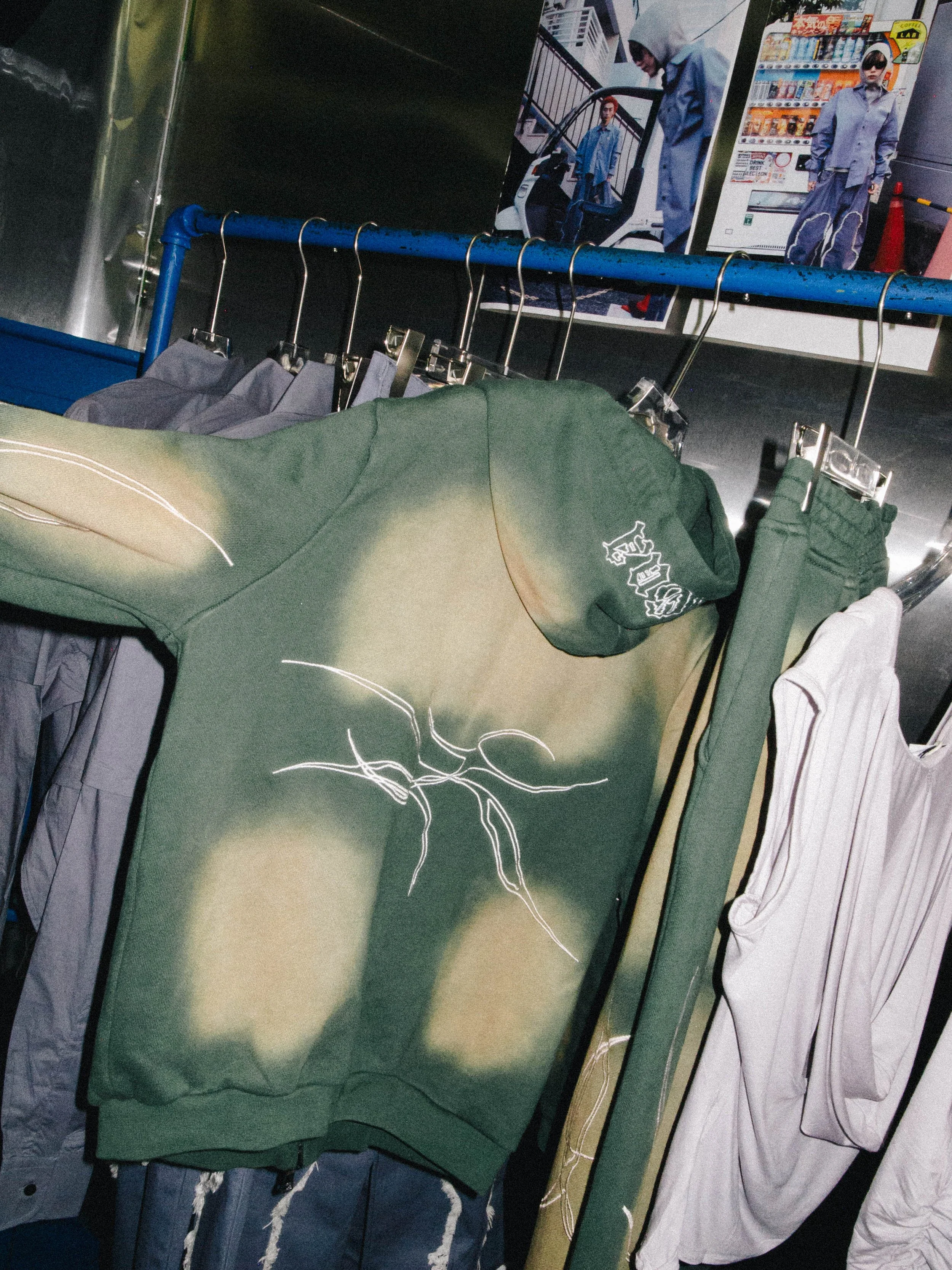 Clothing items hanging on a rack, including a green hoodie with a graphic design, a plastic white shirt, and other garments in the background, with posters on the wall behind.