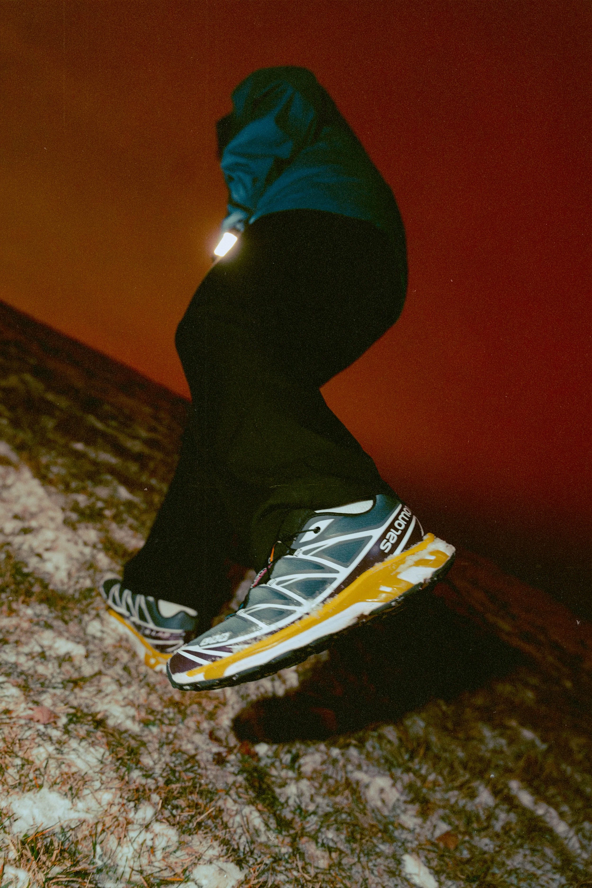 A person wearing a blue jacket, black pants, and Salomon trail running shoes jumping over snow-covered ground at night with a dark red sky.