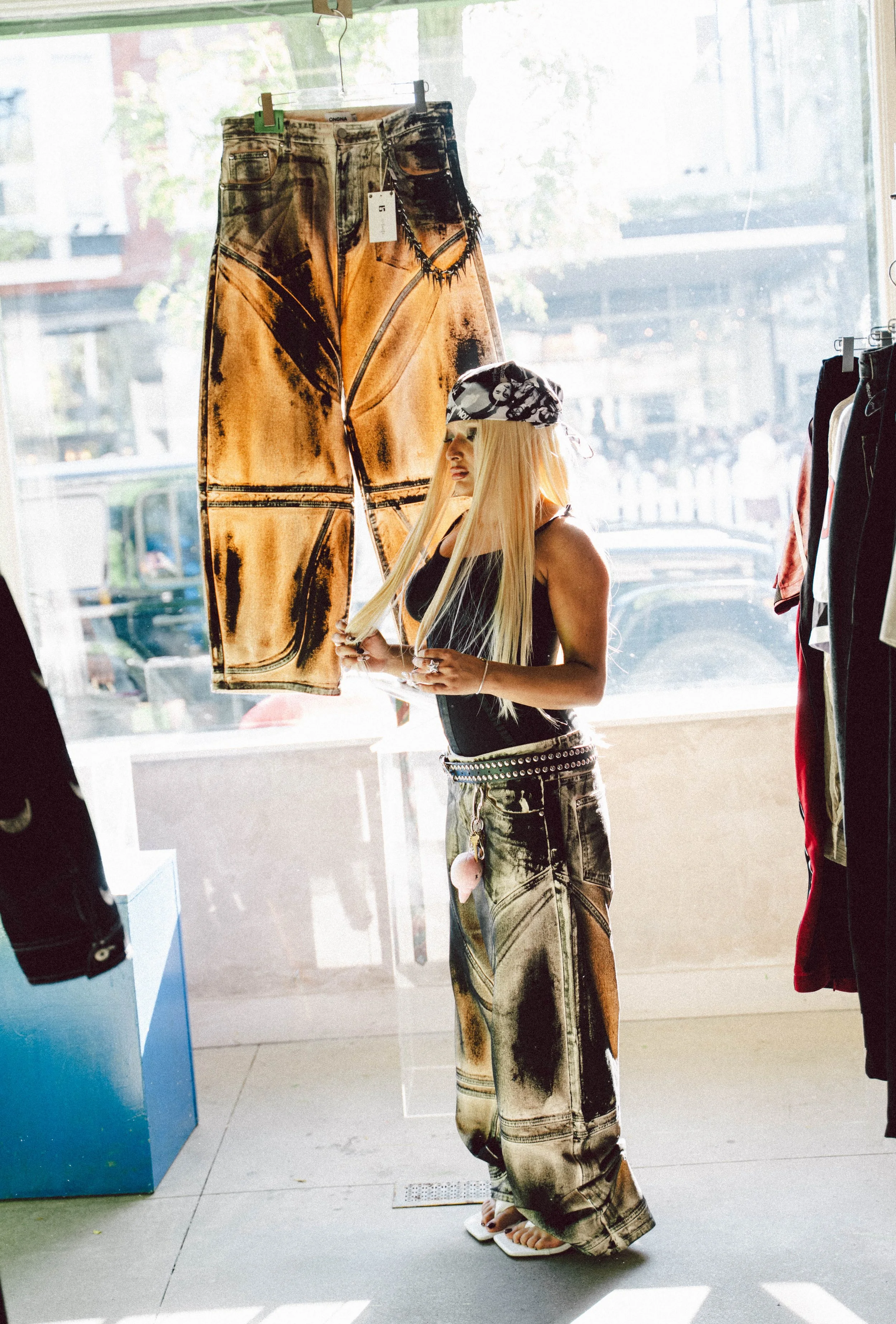 A person with long blonde hair wearing a black tank top, baggy paint-splattered jeans, and a black and white bandana, standing inside a clothing store, holding a piece of clothing while looking out the window.