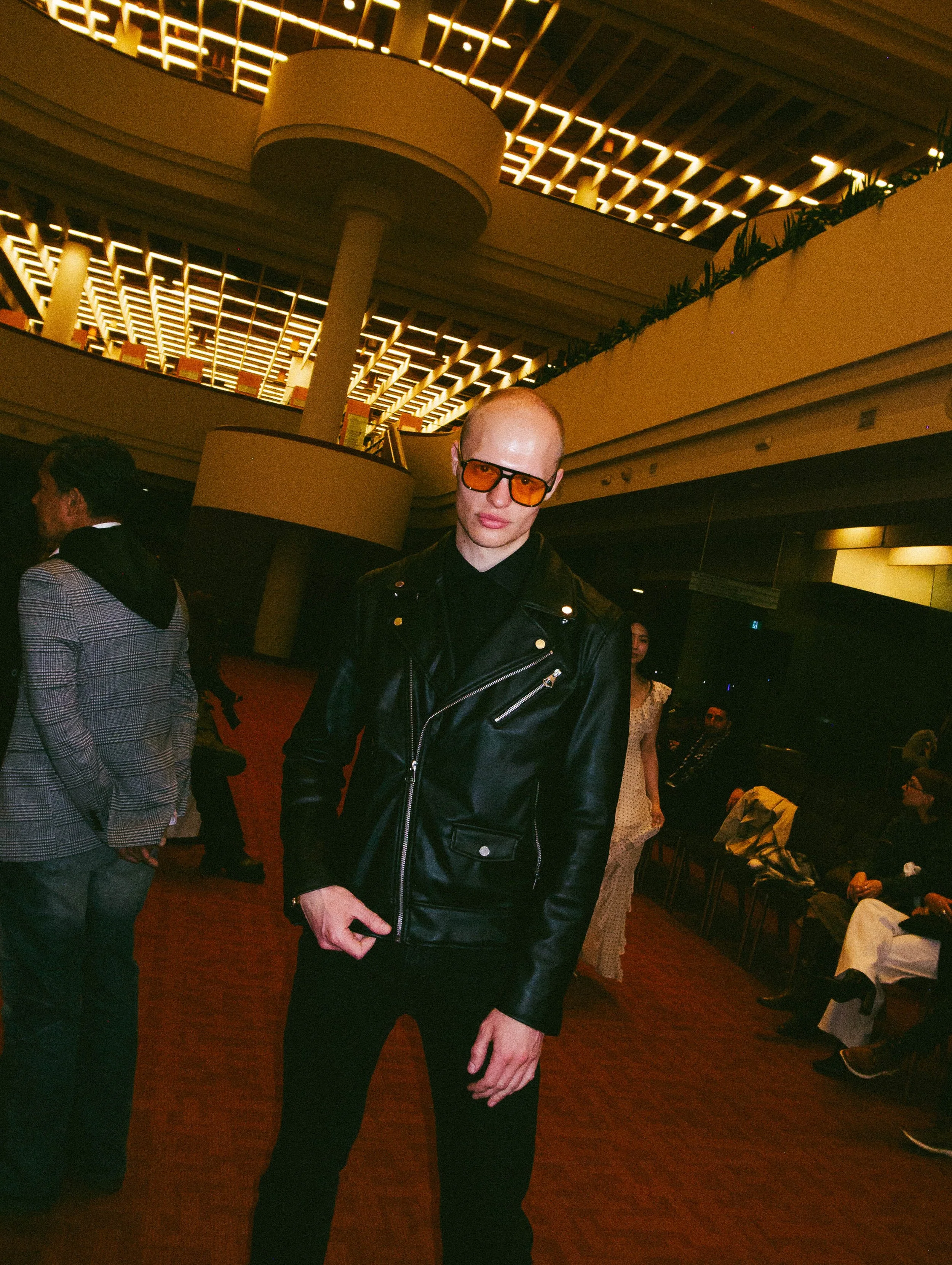 A man wearing a black leather jacket and orange tinted glasses standing in a modern, well-lit interior space with other people seated and standing around.