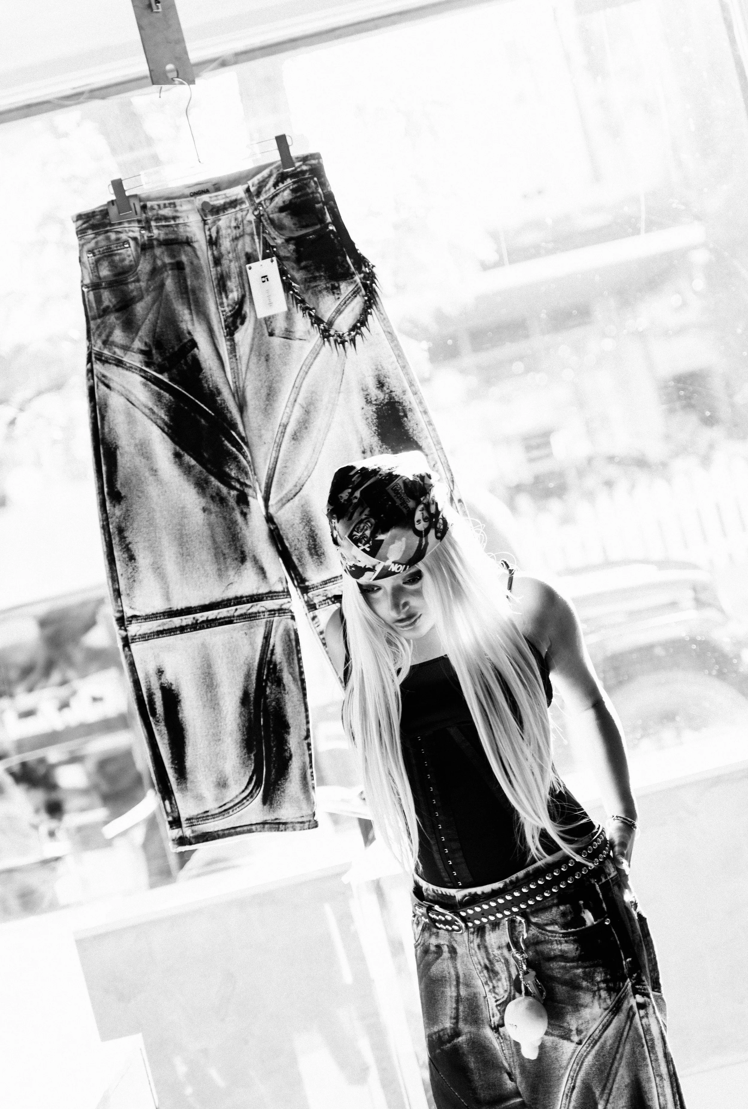 A mannequin with long blonde hair wearing a bandana, black top, and baggy acid-wash jeans, posed indoors with jeans hanging on a rack nearby.