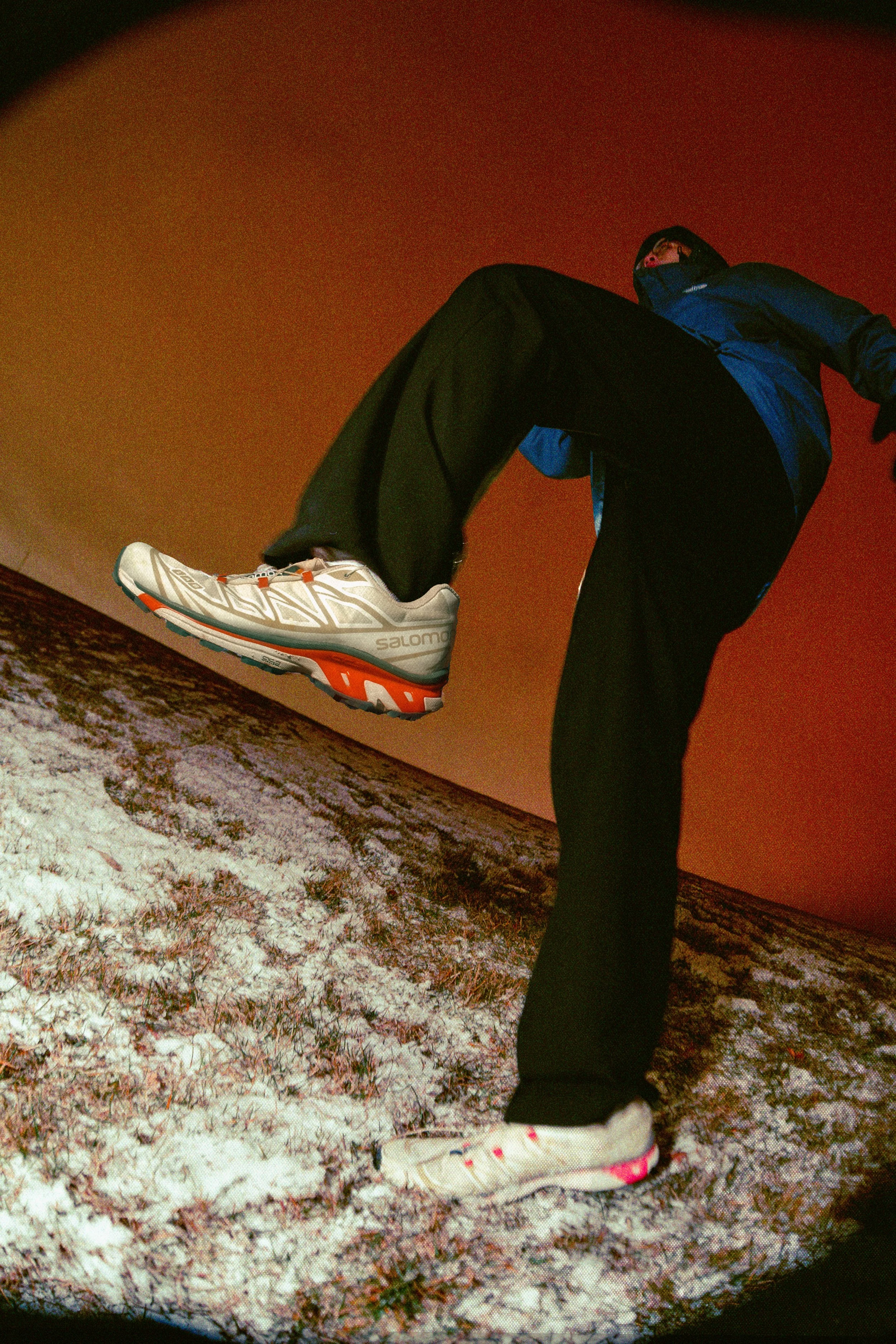 Person wearing athletic shoes and outdoor gear walking on snow-covered ground at dusk or dawn.