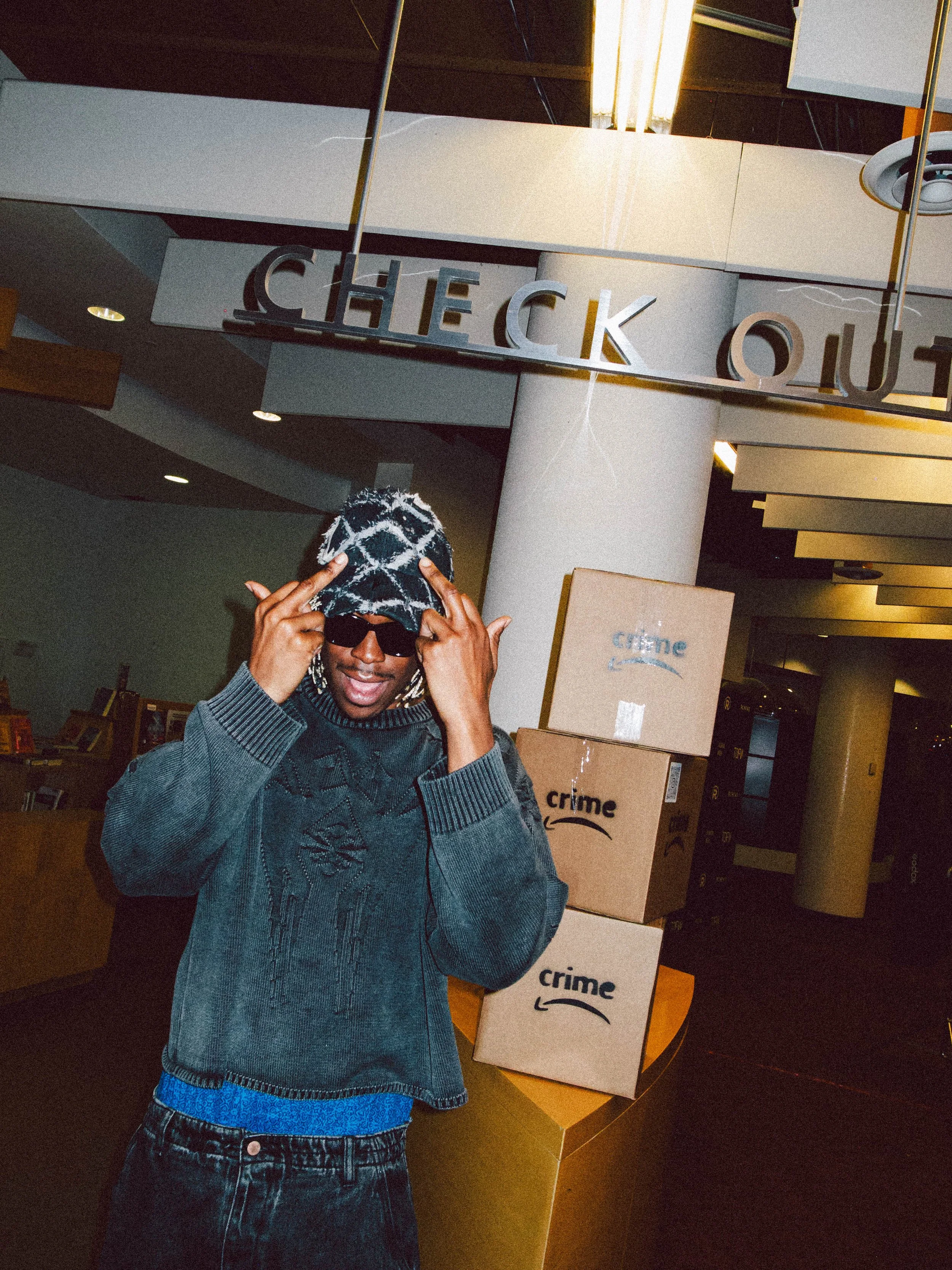 A young man wearing sunglasses, a patterned hoodie, and jeans, standing at an airport check-in area with stacked boxes labeled 'crime' behind him.