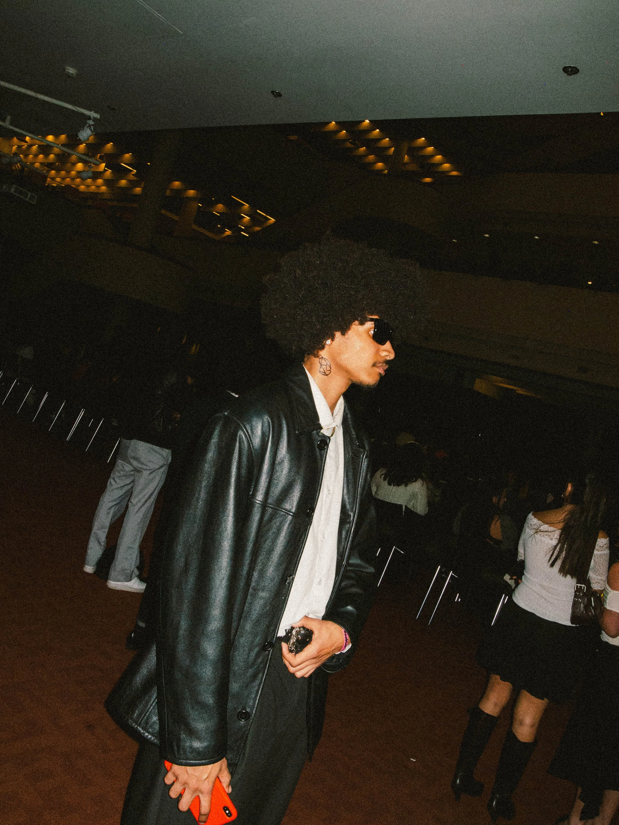 A young man with an afro hairstyle wearing sunglasses, a black leather jacket, and a white shirt, standing indoors at an event or party, holding an orange phone in his left hand.