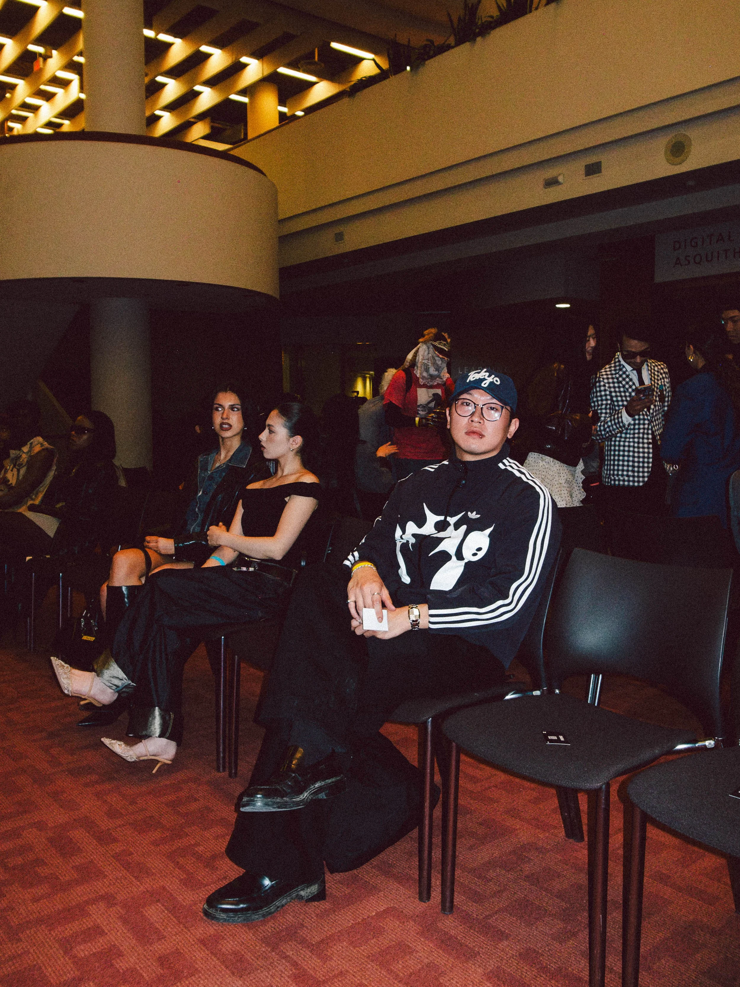 People sitting in a theater or conference hall, with a man in the foreground wearing glasses, a black and white jacket, and a cap, and two women behind him dressed in dark outfits. Others are standing in the background, some using their phones.