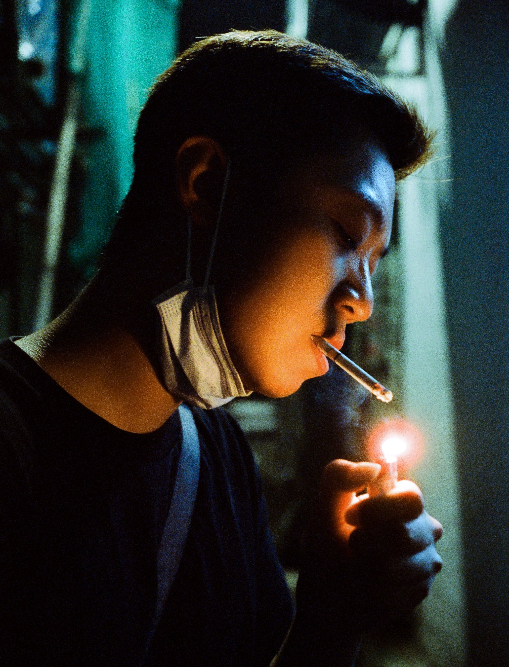 A young man with dark hair lighting a cigarette with a match, wearing a face mask around his neck and headphones, in a dimly lit environment. that shot with 35mm film that call cinestill 800t 