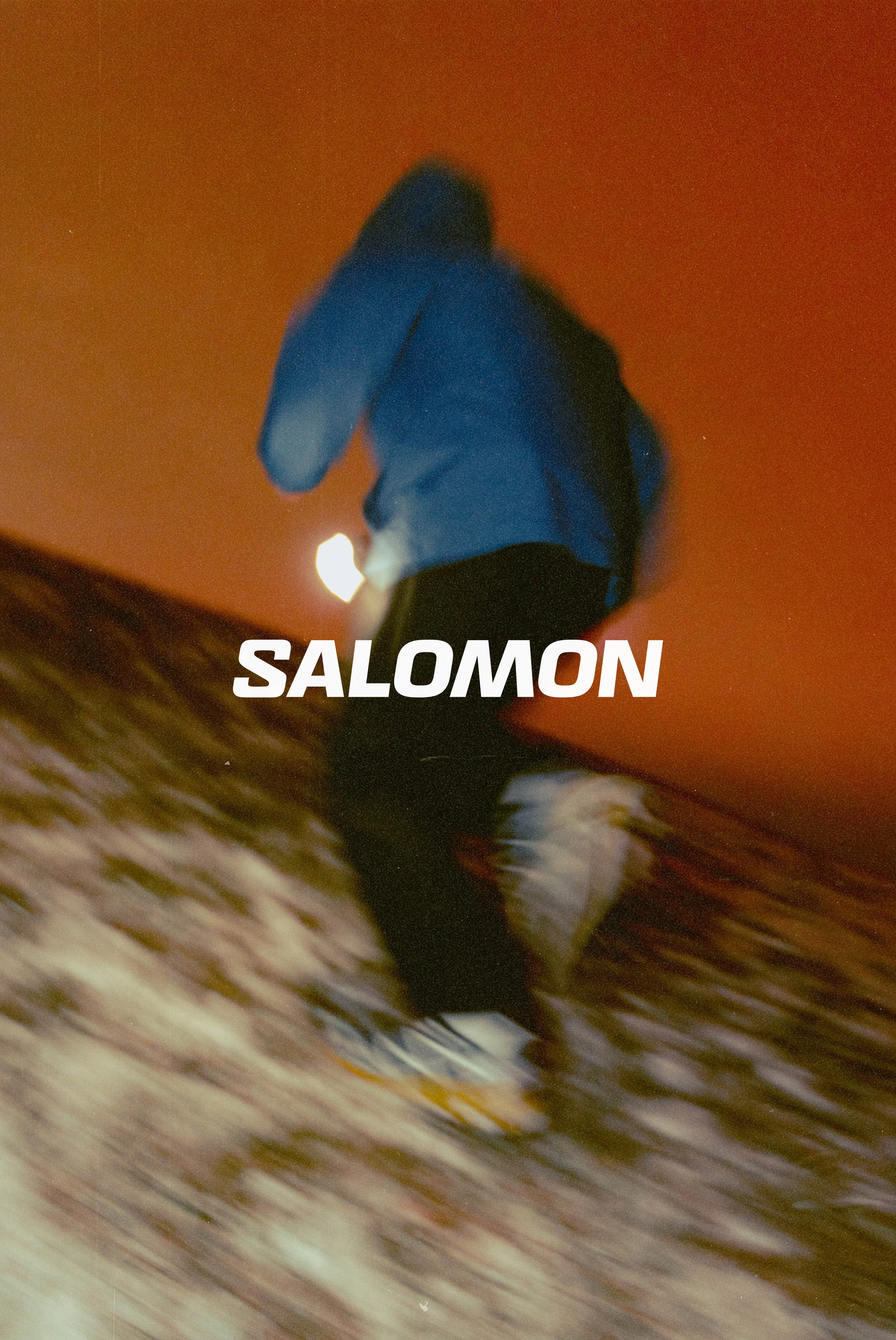 A person wearing a blue hoodie and black pants walking on a wet surface at night with a bright streetlight and a red sky in the background. The word 'SALOMON' is overlaid on the image.