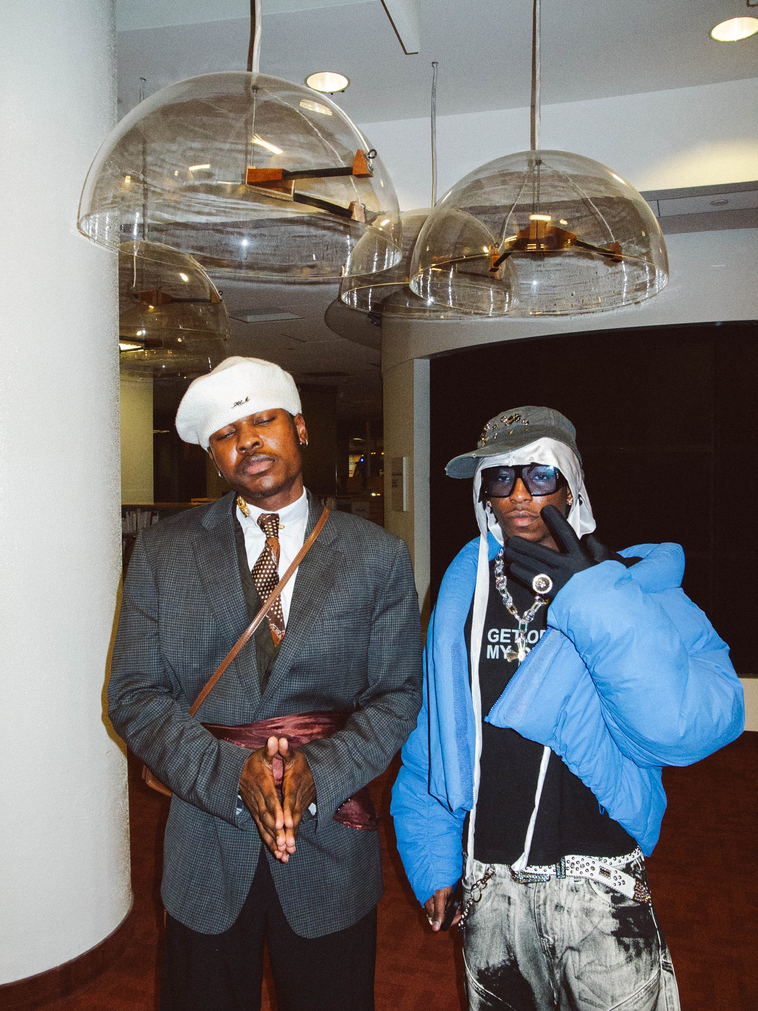 Two men standing in an indoor space under modern ceiling lights. One is dressed in a suit with a white beret, and the other is in casual streetwear with sunglasses, a hat, and a blue jacket.