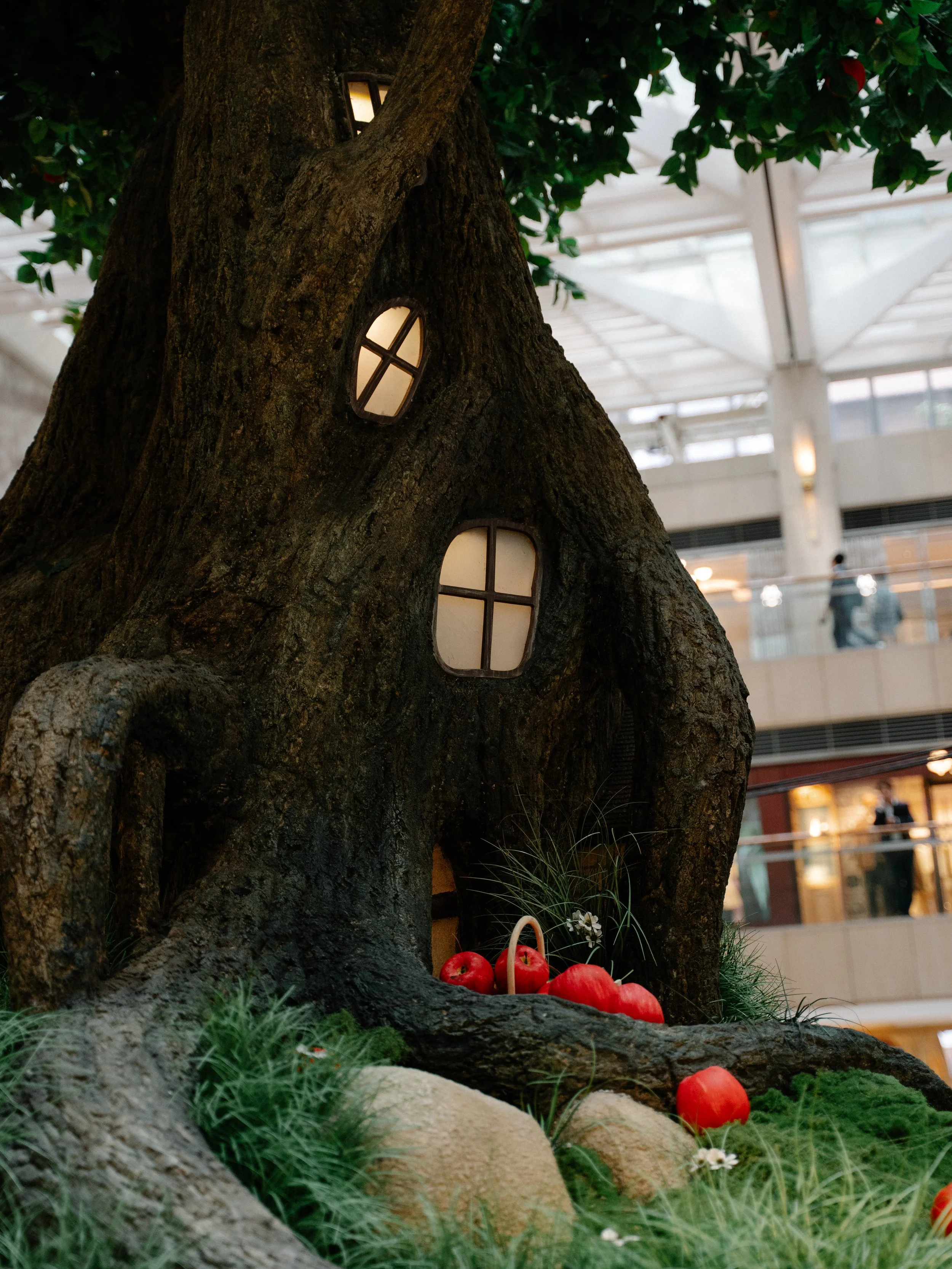 Tree with small house-like windows, decorated with red pumpkins, green plants, and rocks at the base inside a shopping mall.
