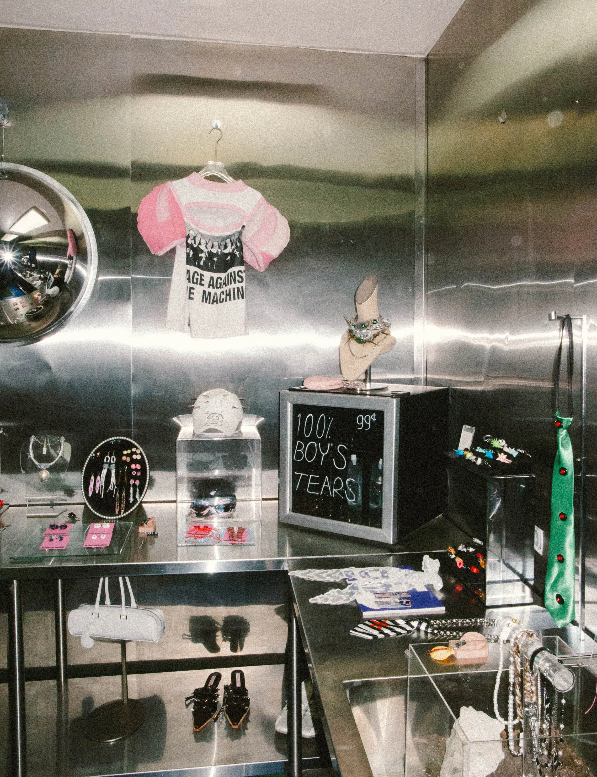 A boutique display with jewelry, accessories, and clothing items on stainless steel surfaces and a chalkboard sign that reads "100% Boy's Tears."