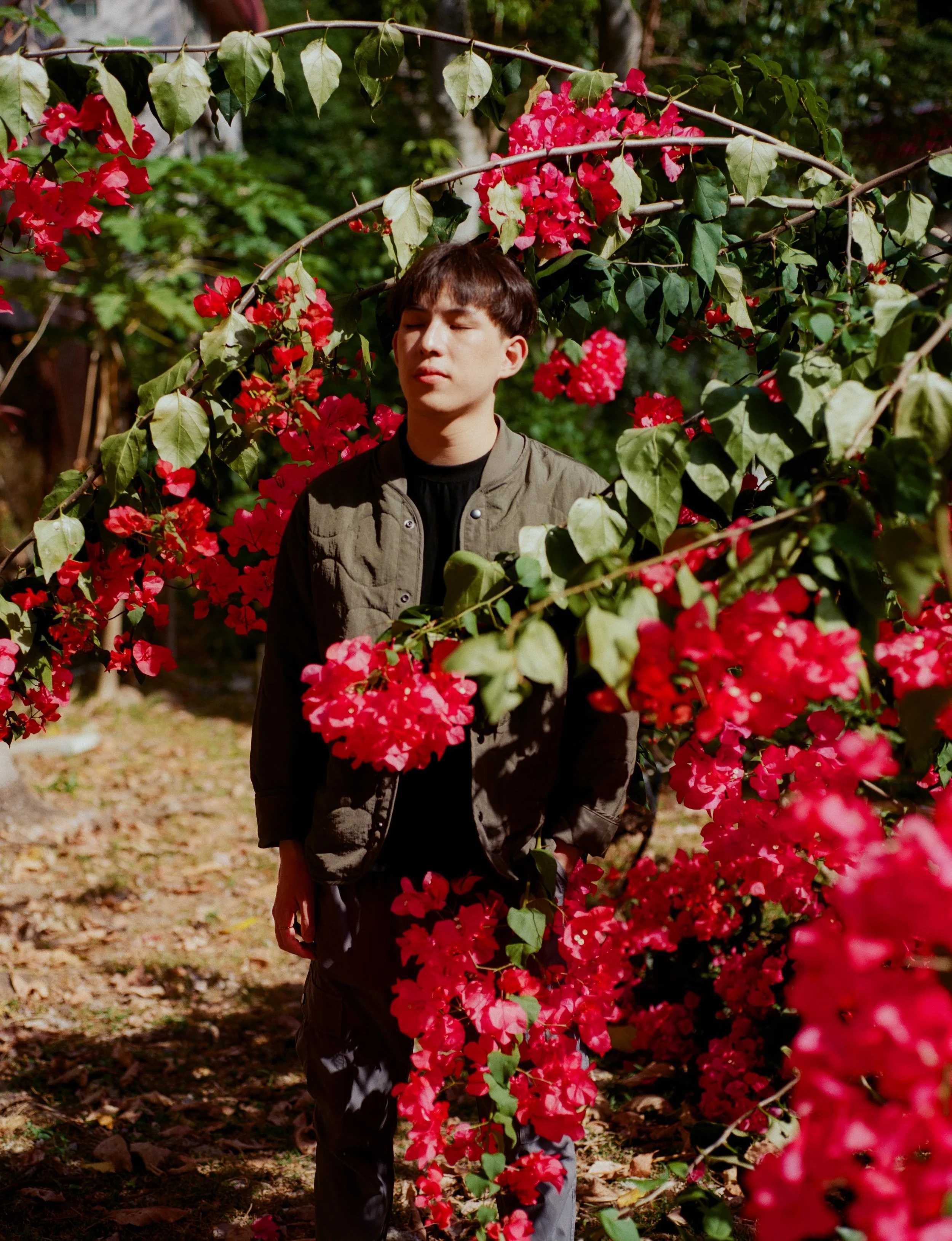 A young man with closed eyes standing among bright pink and red bougainvillea flowers in a garden.