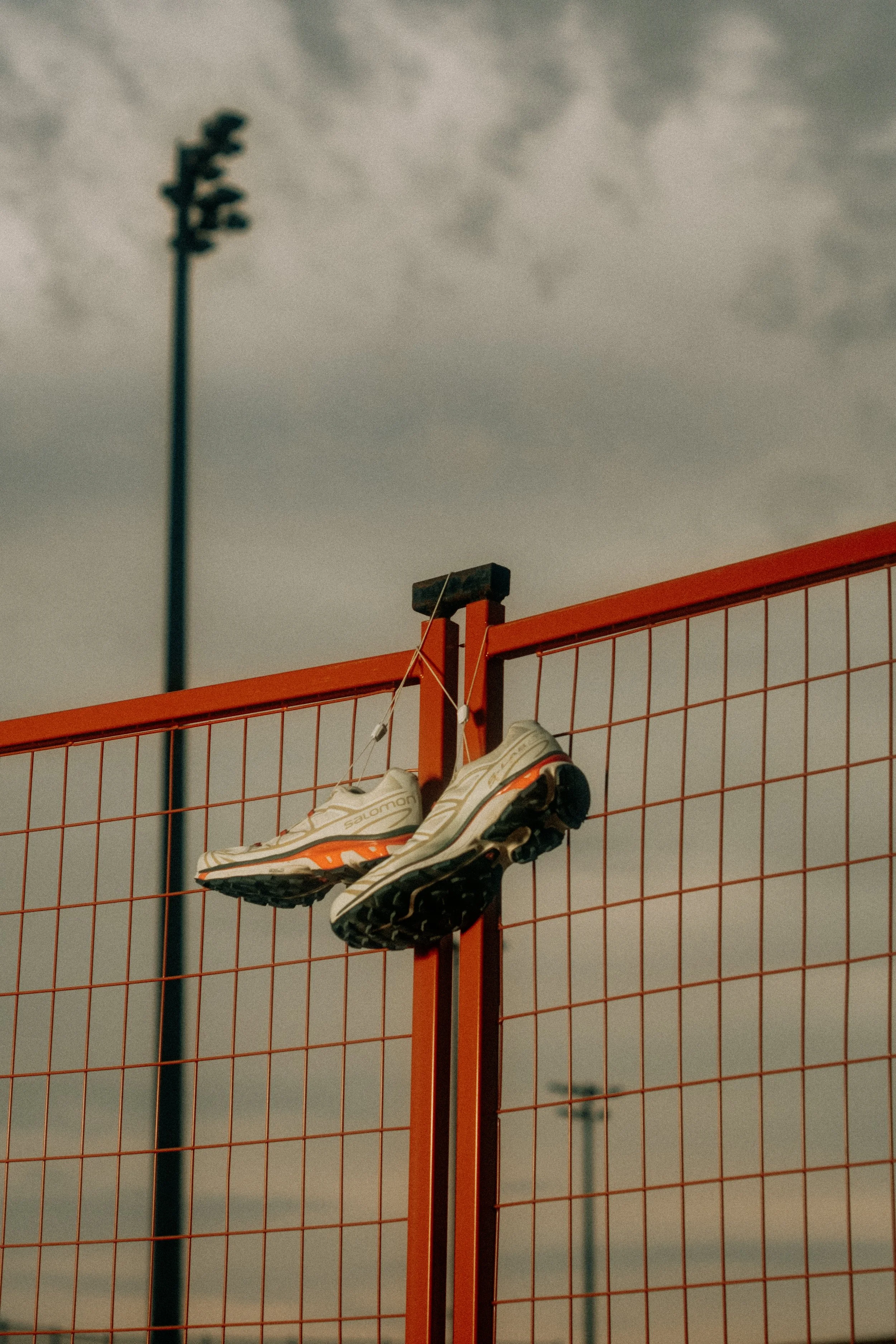 A pair of Salomon xt6 running shoes hanging on a red wire fence with a cloudy sky in the background.