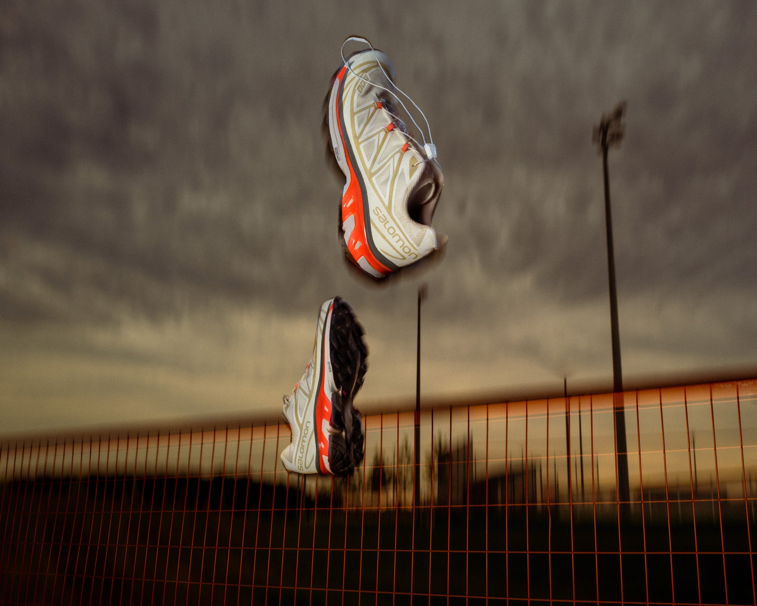 A pair of athletic running shoes "salomon xt6" hanging in the air above a fence during a sunset or dusk, with a cloudy sky and a lamppost in the background.