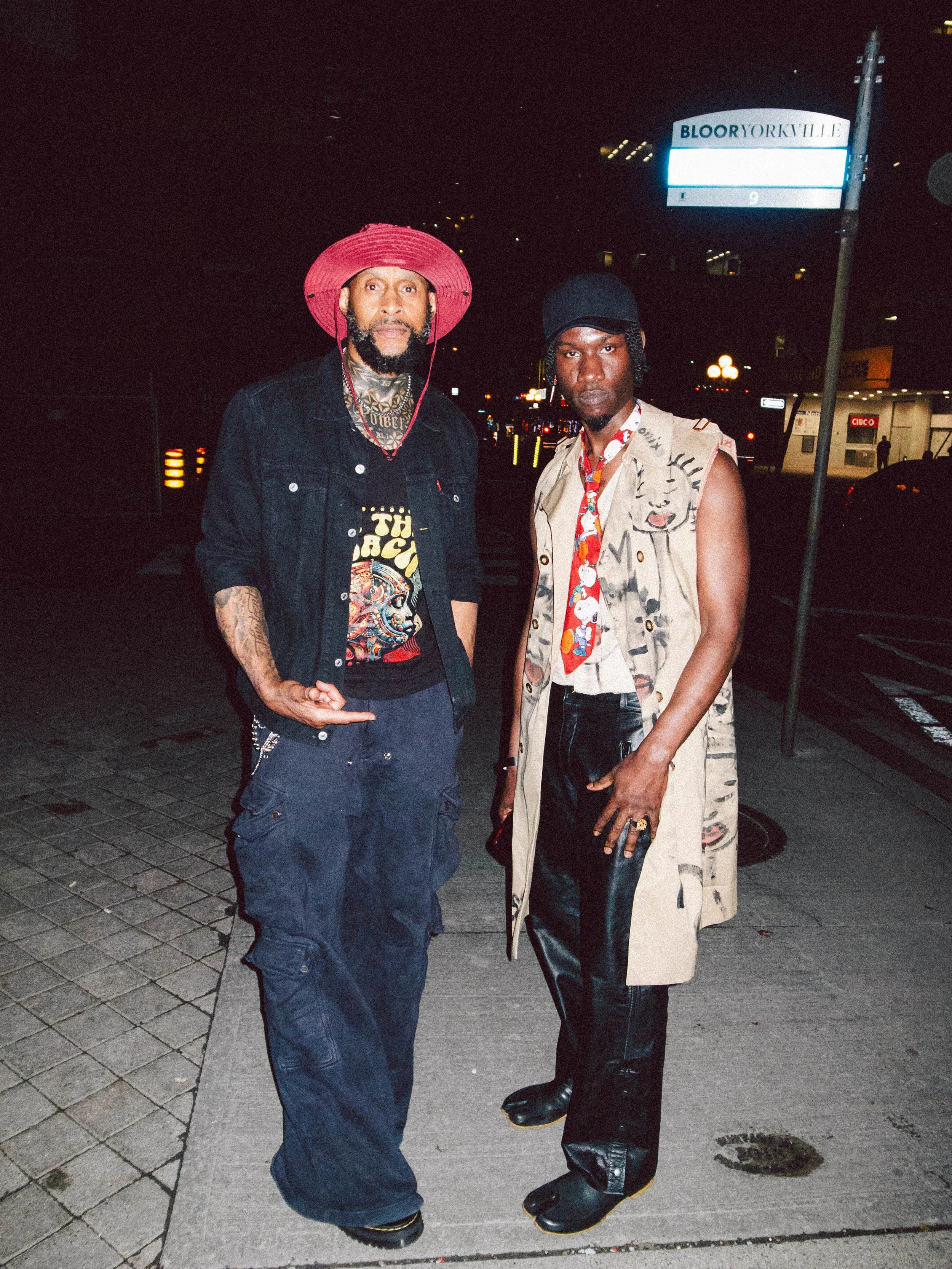 Two men standing on a city sidewalk at night, one wearing a red wide-brimmed hat, black denim jacket, and dark pants, the other wearing a bucket hat, sleeveless patterned coat, and shiny black pants.