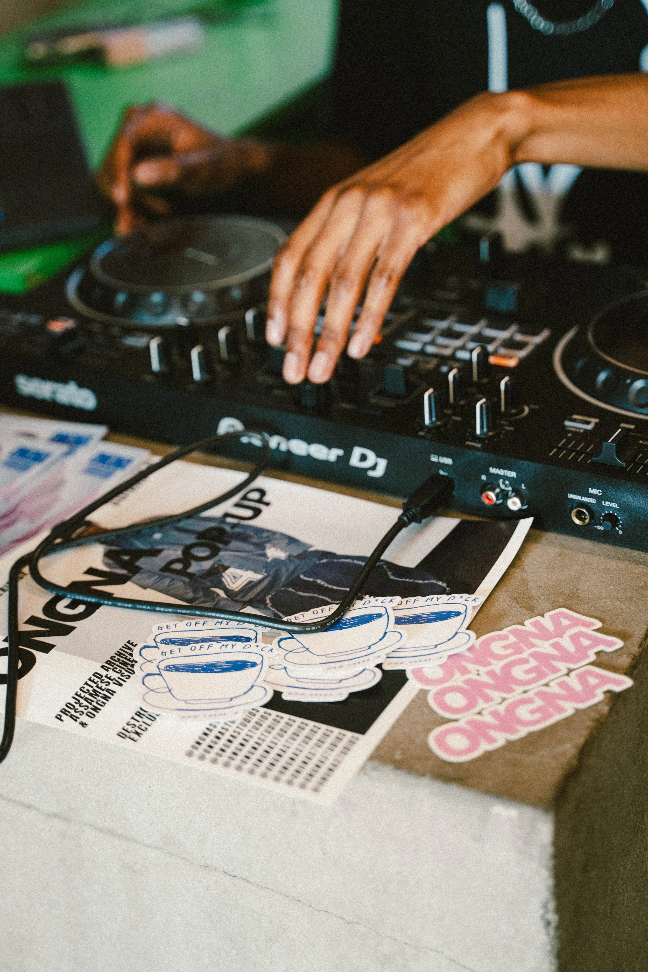 Close-up of a DJ's hand operating a Pioneer DJ controller at a music event, with stickers and a flyer on the table.