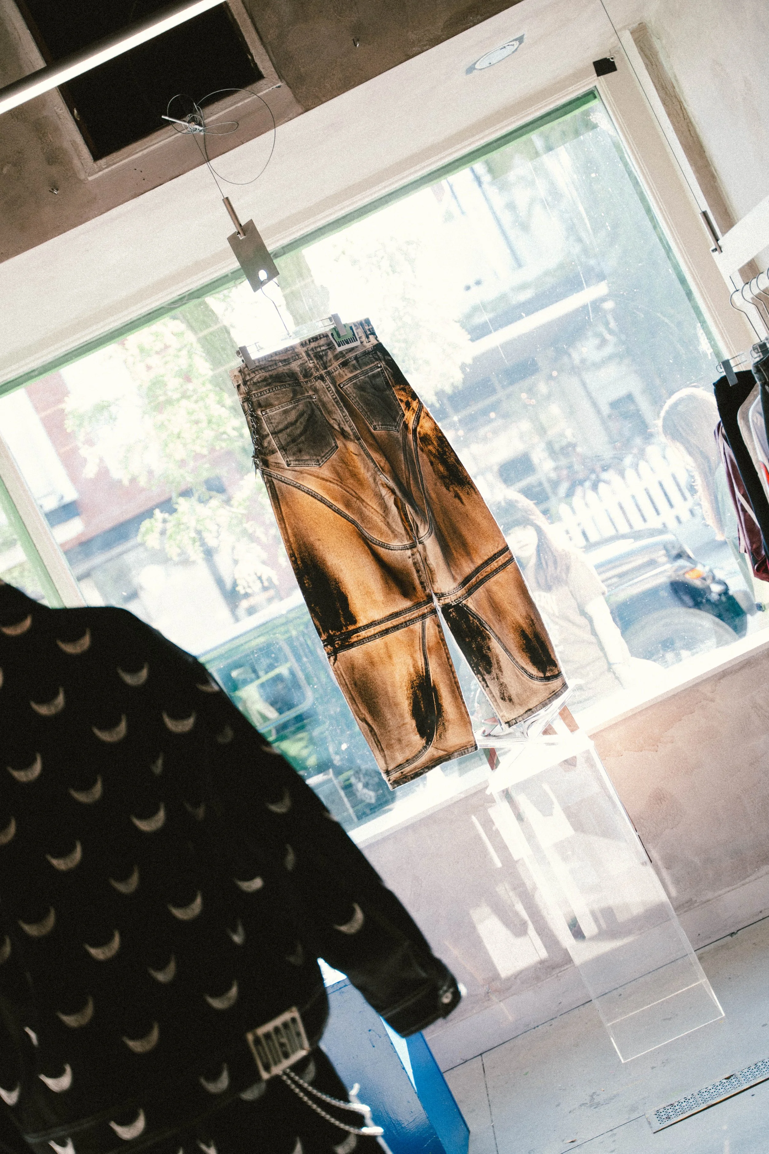 A pair of pants with a tie-dye design hanging in a store window, with reflections of people and trees outside.