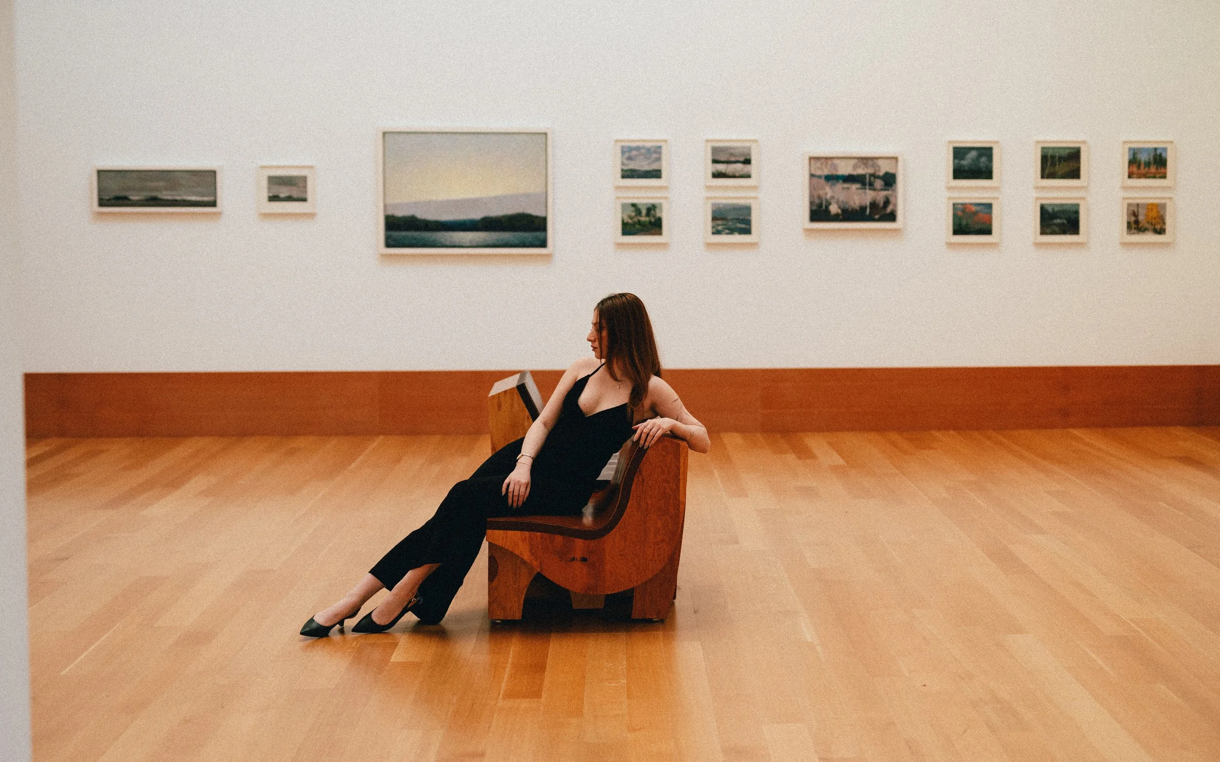A woman in a black dress sitting on a wooden bench in an art gallery with framed paintings on the white wall behind her.