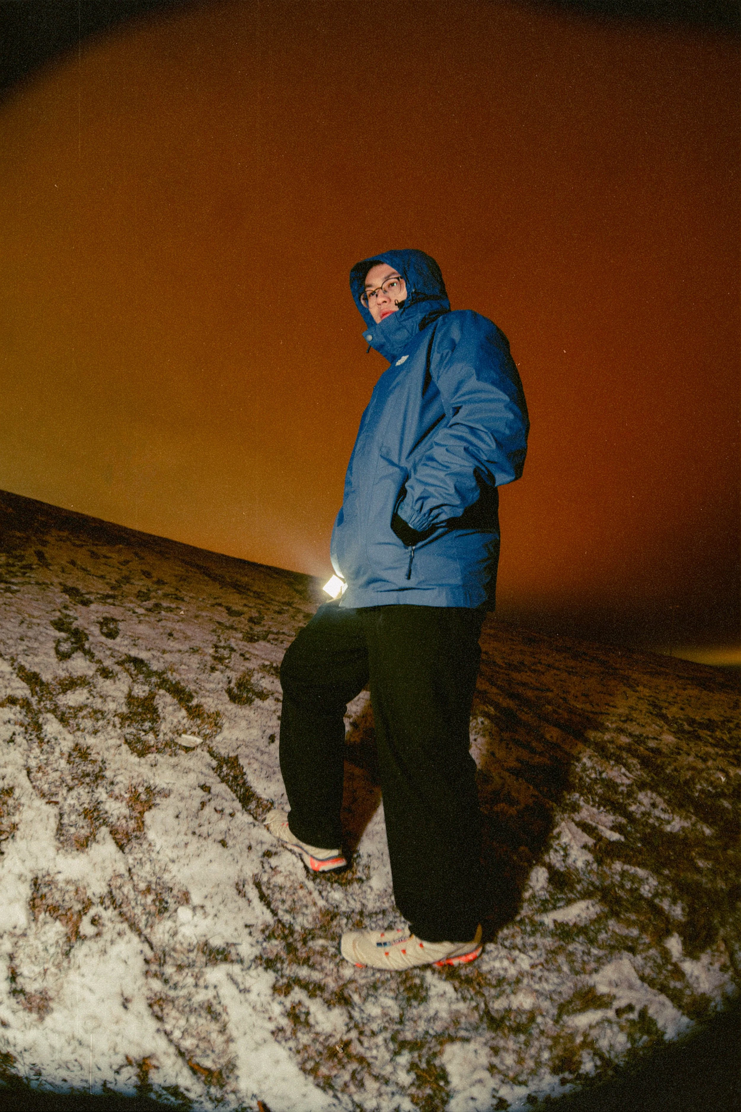 A person in a blue winter jacket, black pants, and white sneakers stands on a snowy slope at night, with a reddish-orange sky in the background.