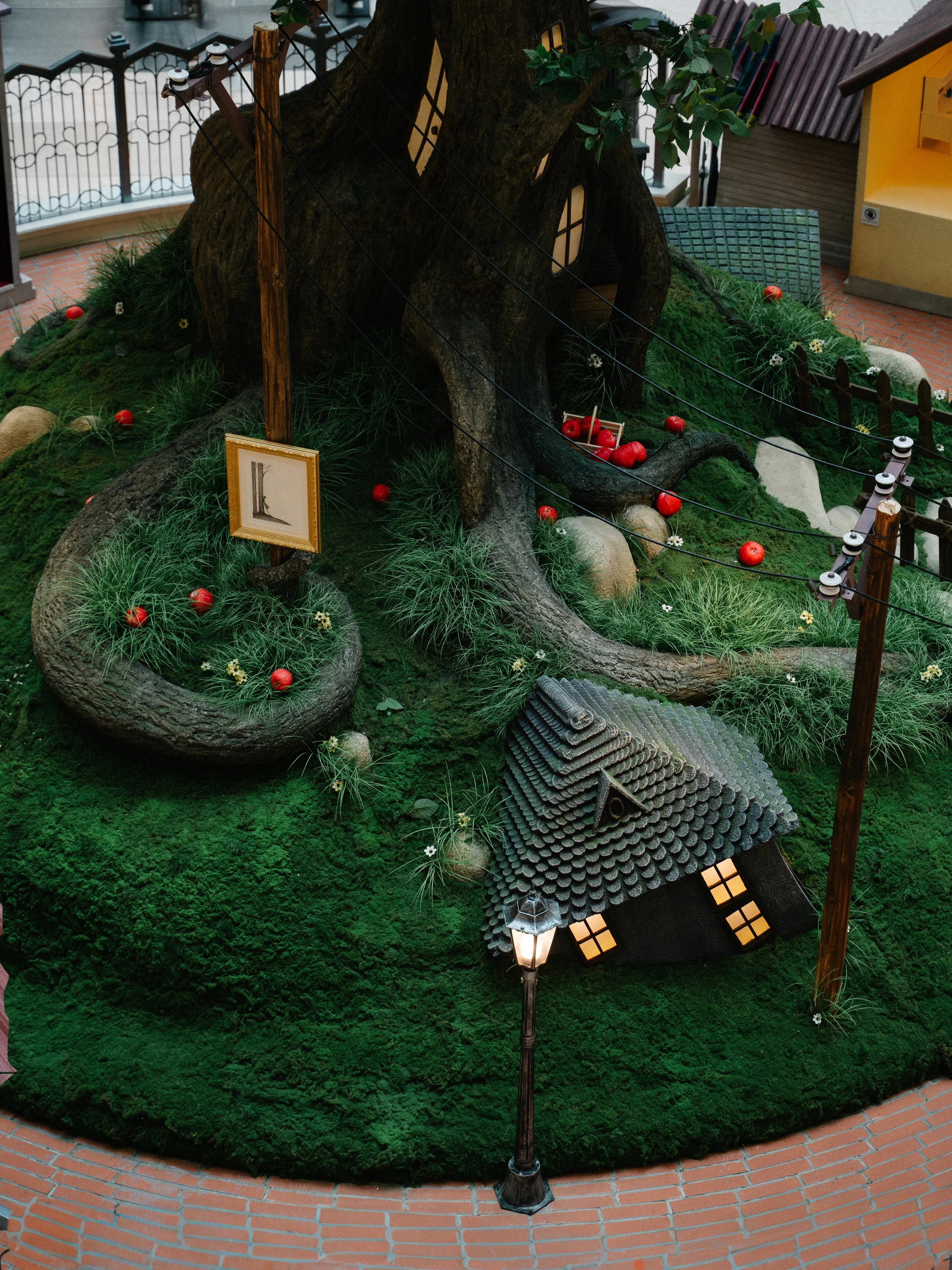 Large artificial tree with a hollowed trunk and small windows, surrounded by green moss, grass, and scattered red apples, with a small house with a steep tiled roof and glowing windows nearby. There are power lines, wooden poles, a street lamp, and a