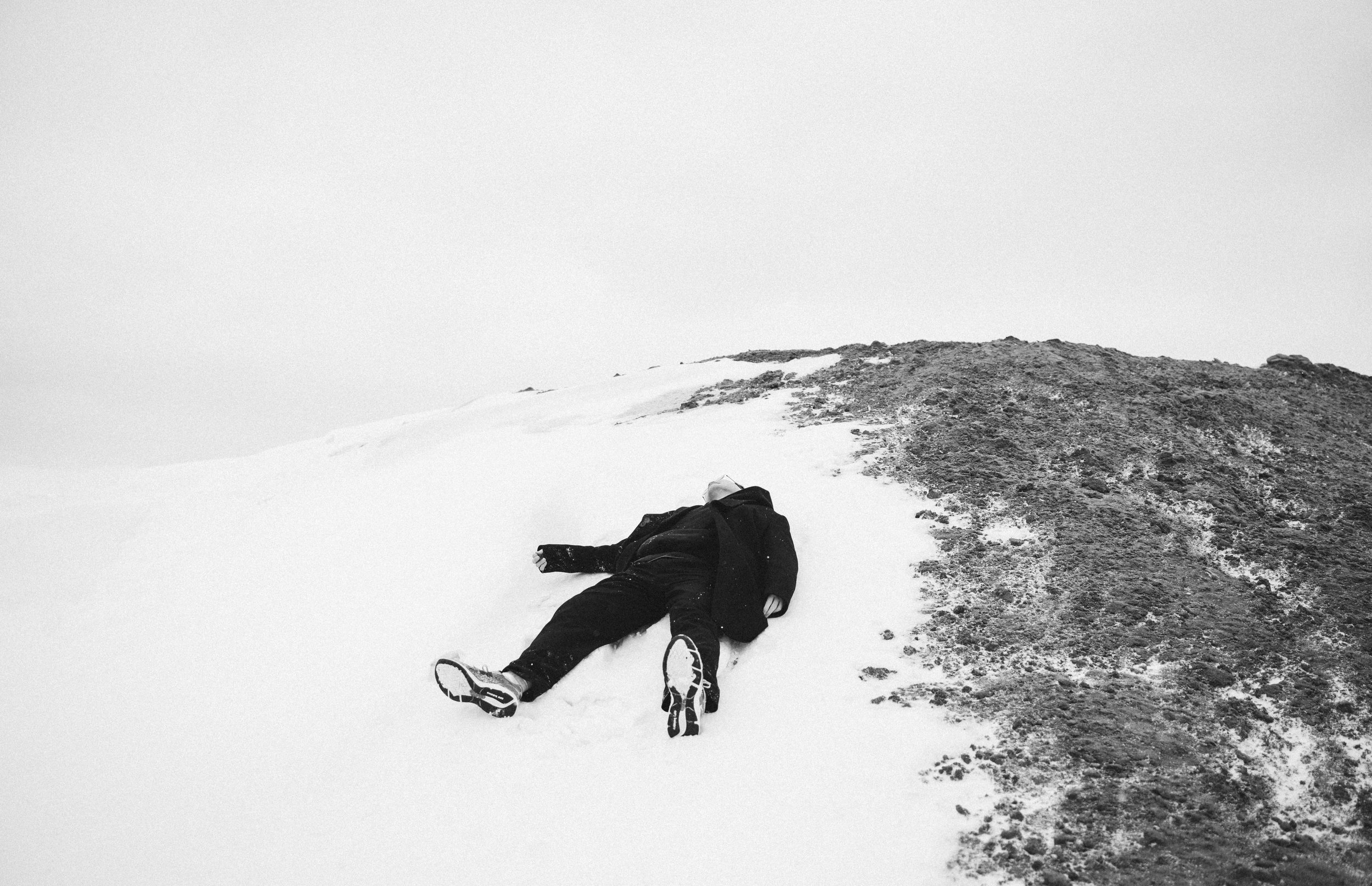 Person lying on snow-covered ground near a rocky hillside on a cloudy day.