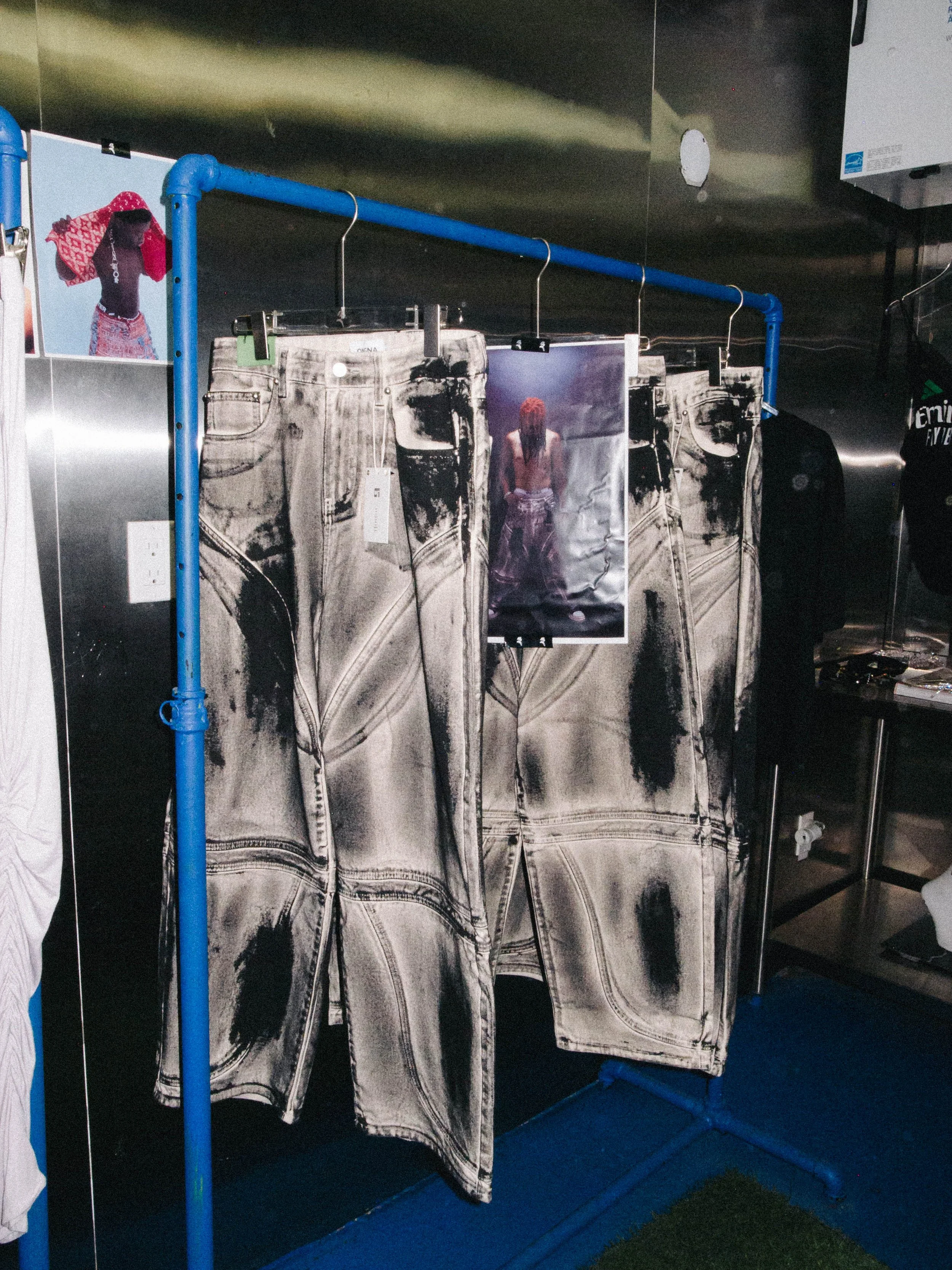 Pair of jeans with black and white tie-dye pattern hanging on a blue clothes rack, with a poster of a person with long hair and bare back hanging in front of the jeans.