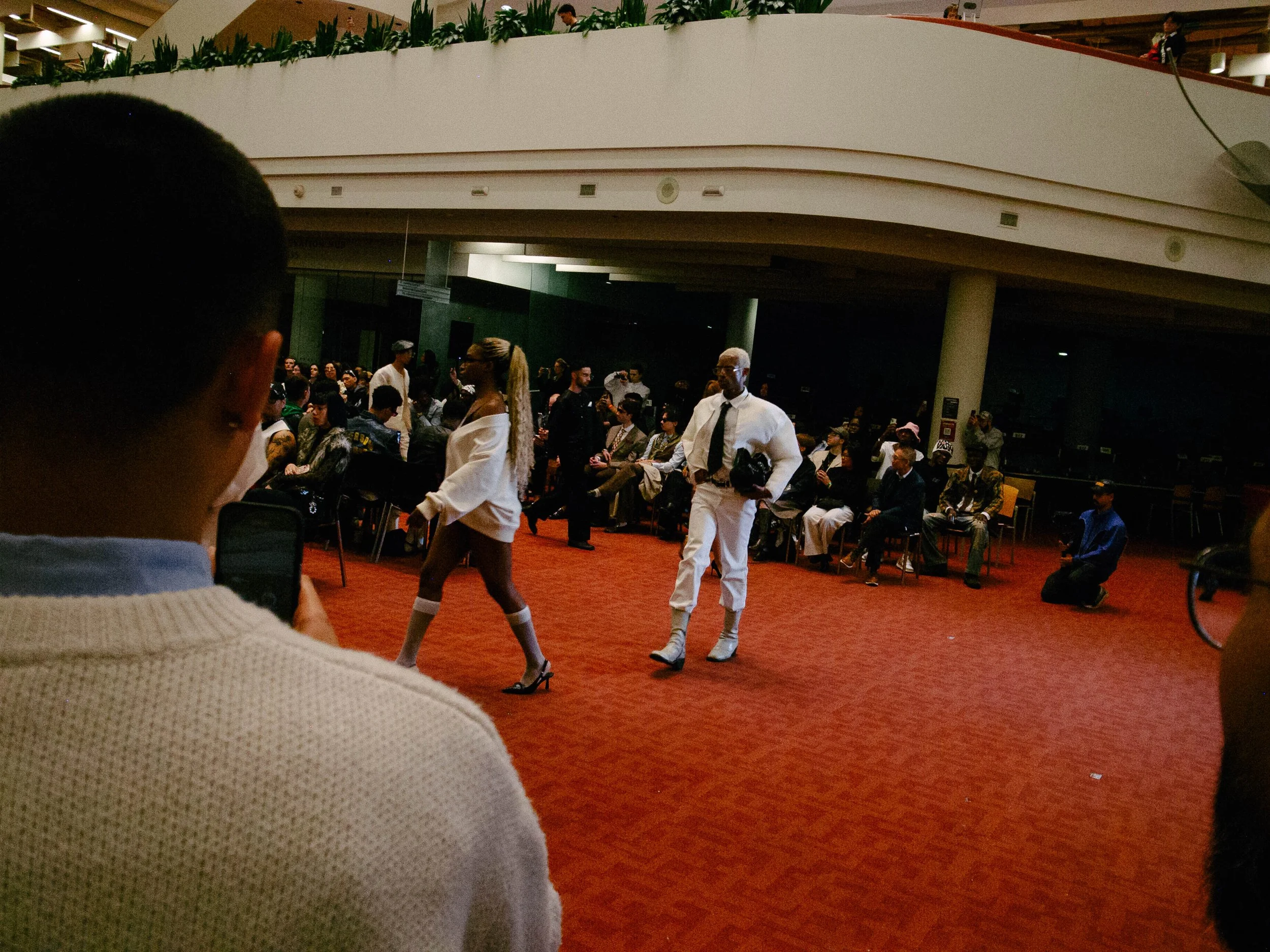 People sitting and watching a fashion show in an indoor venue with a red carpet floor, some models walking on the runway, and individuals in the audience taking photos.