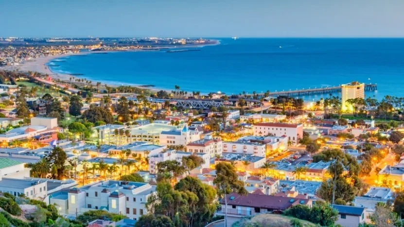 Brightly lit Ventura coastal cityscape with a beach, pier, and ocean in the background at dusk.
