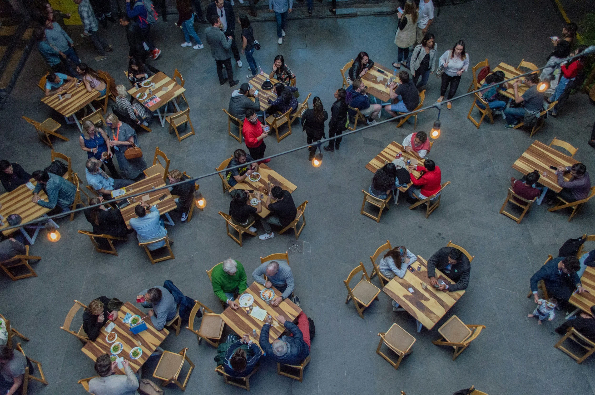 People dining at tables in a busy restaurant or cafe during the evening, with some waiting in line and others eating.