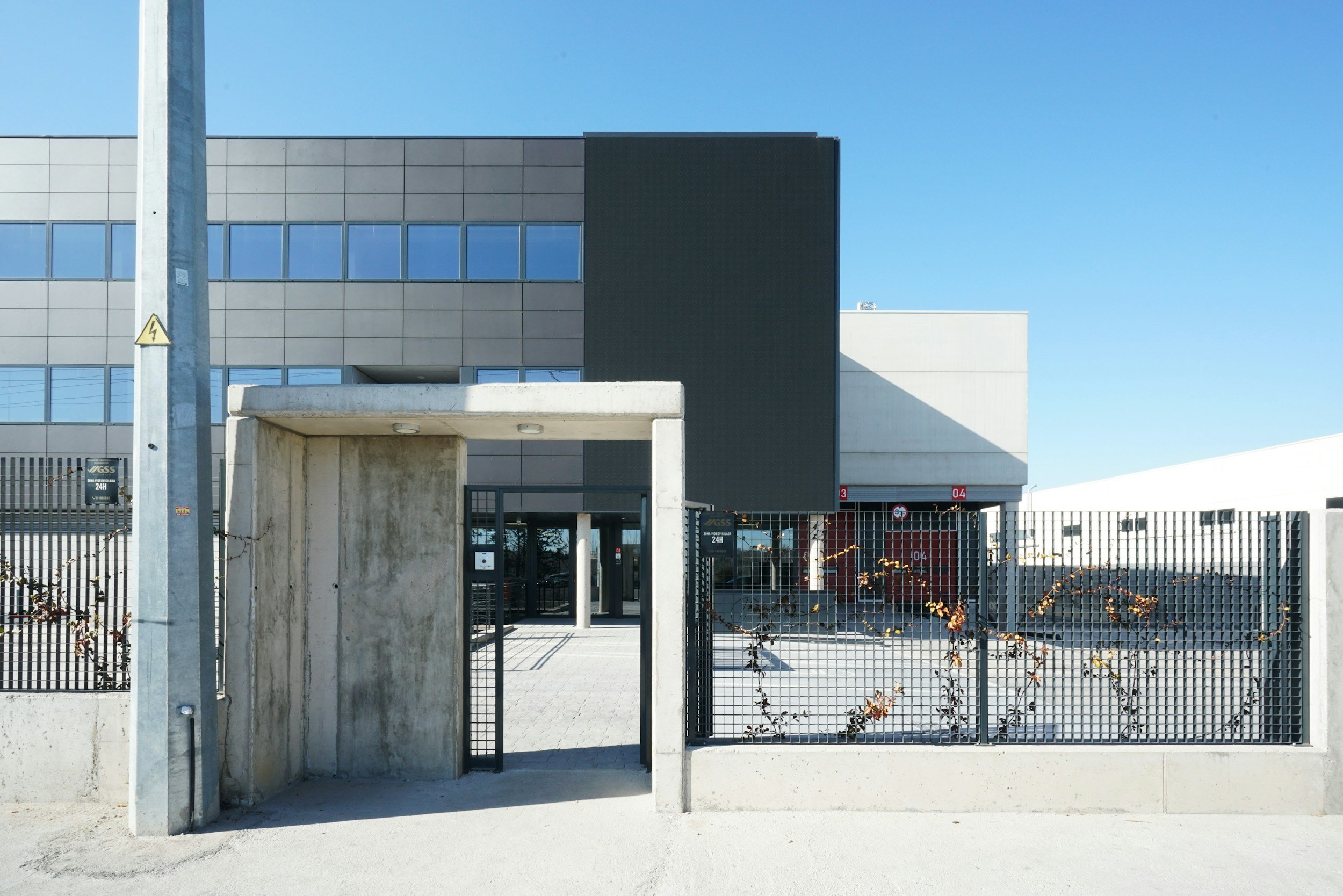Modern multi-story industrial building with large windows, gated entrance, and clear blue sky.