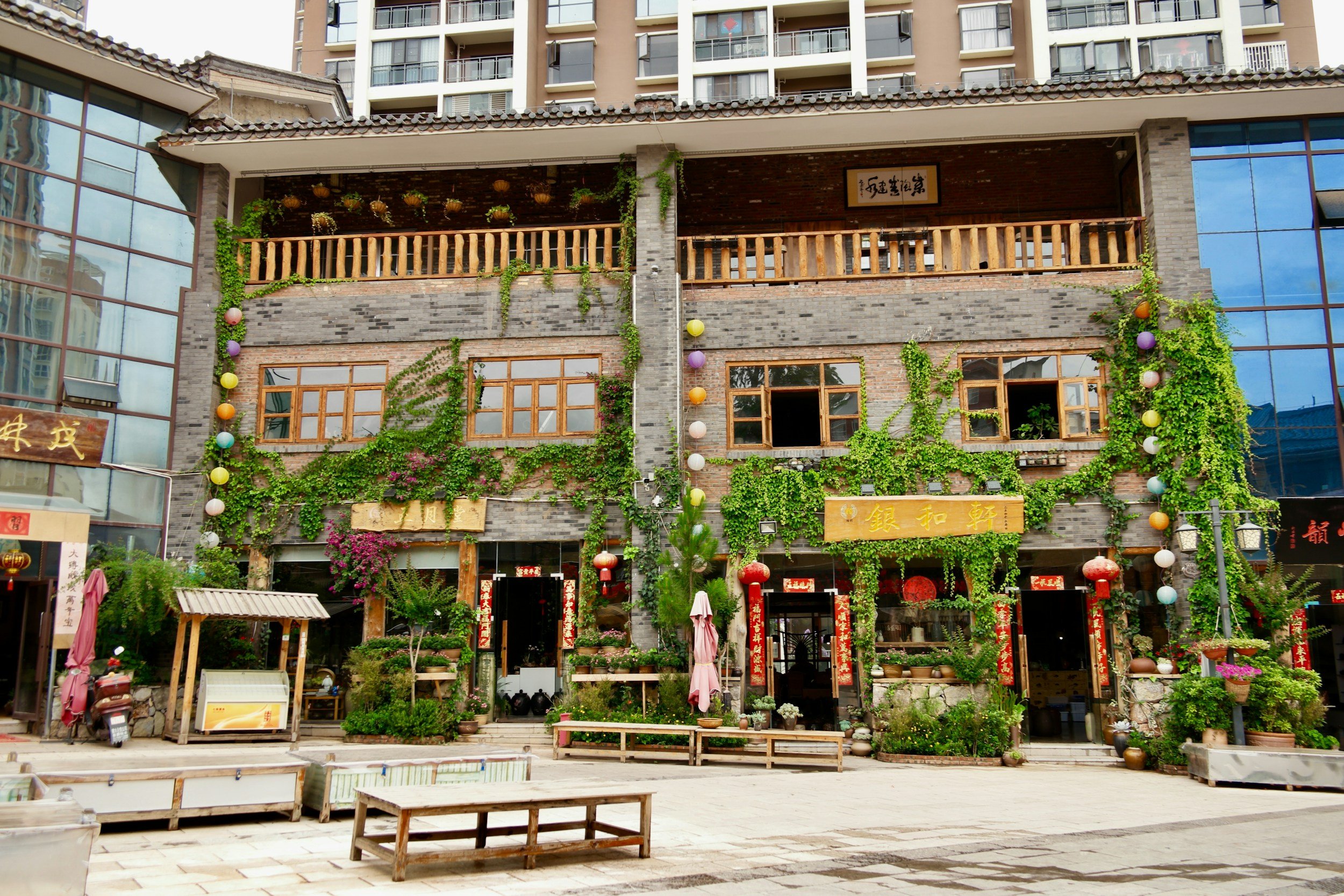 Front view of a rustic multi-story retail building with brick and wooden accents, decorated with green vines and colorful hanging lanterns, located in an urban area with high-rise buildings in the background.