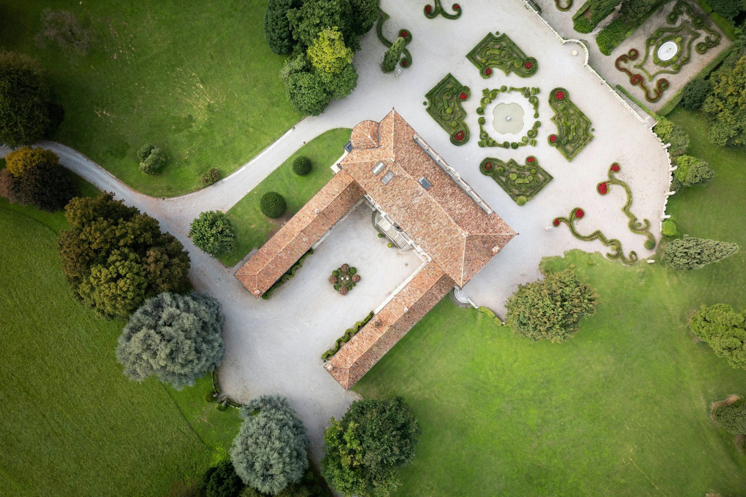 An aerial view of a large estate with a primary building with a terracotta tile roof, surrounded by well-manicured gardens, trees, and gravel pathways. There are intricate garden designs with trimmed hedges and red flowers.