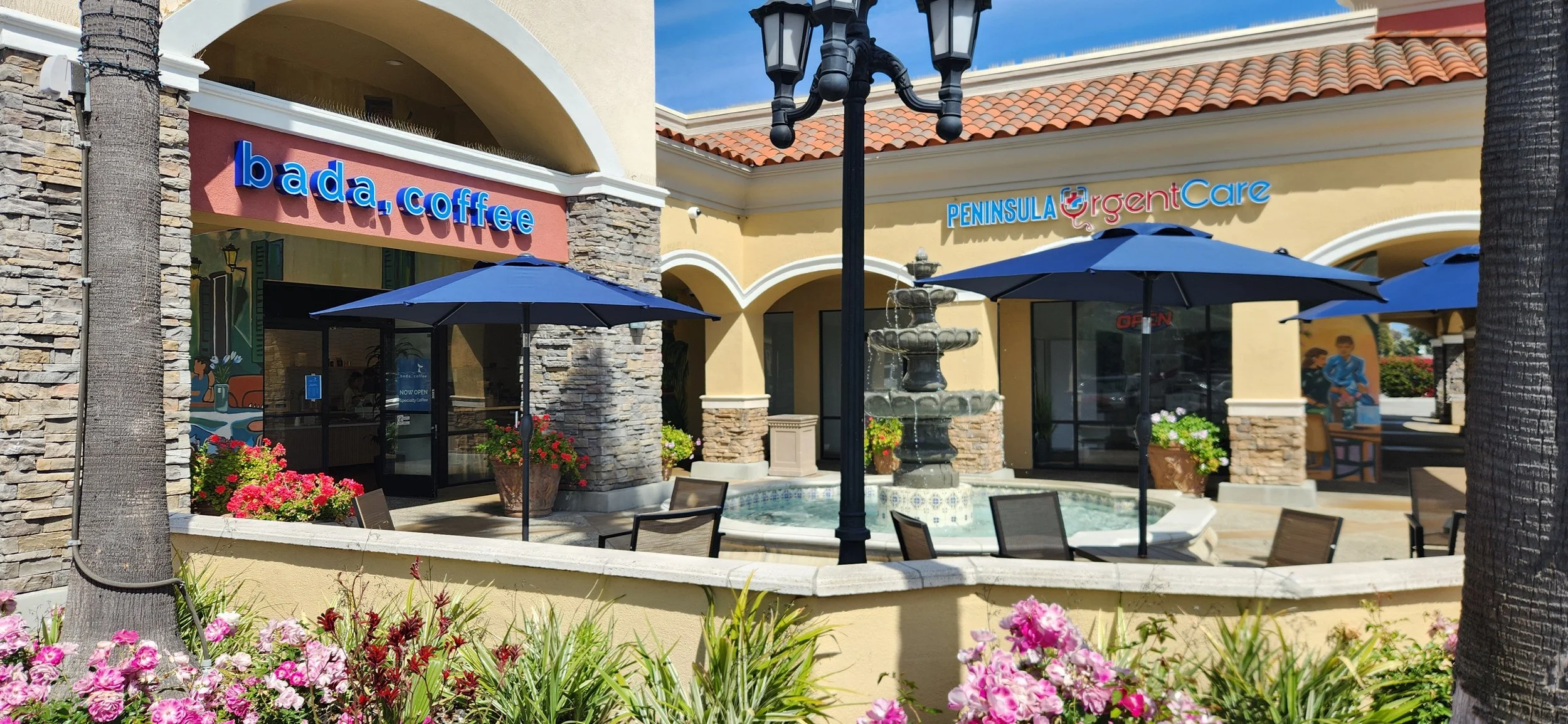 Outdoor shopping plaza with storefronts labeled 'bada, coffee' and 'PENINSULA Urgent Care', featuring a fountain, patio tables with umbrellas, palm trees, and flowering plants.