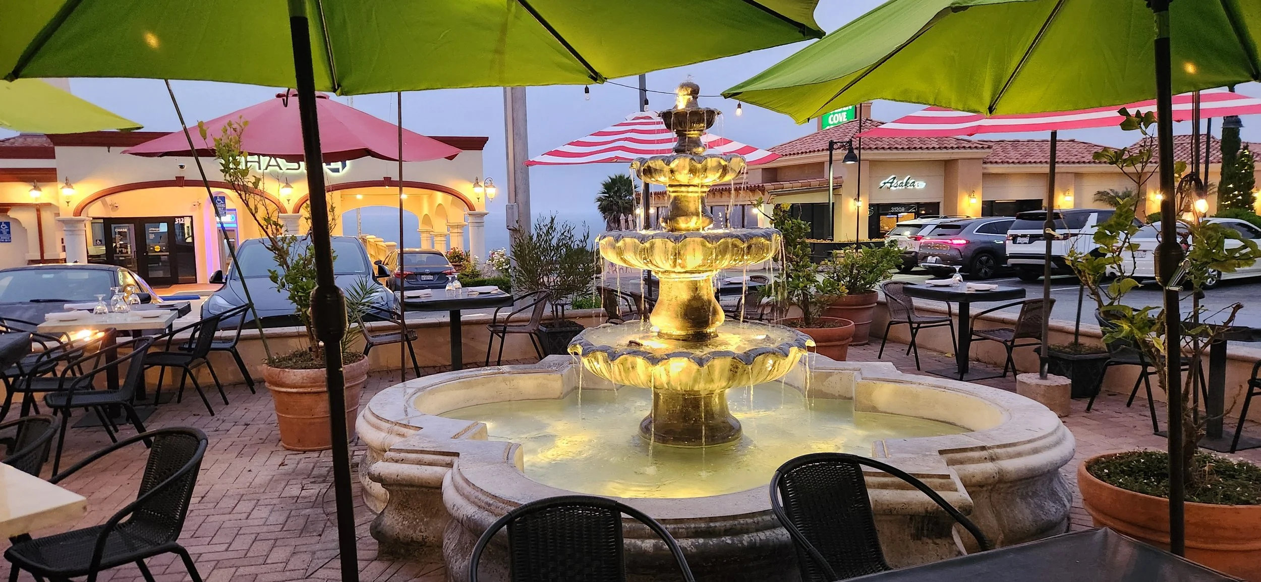 Outdoor restaurant patio with a central illuminated fountain, surrounded by tables and chairs under large umbrellas, parked cars in the background, and buildings with retail storefronts.
