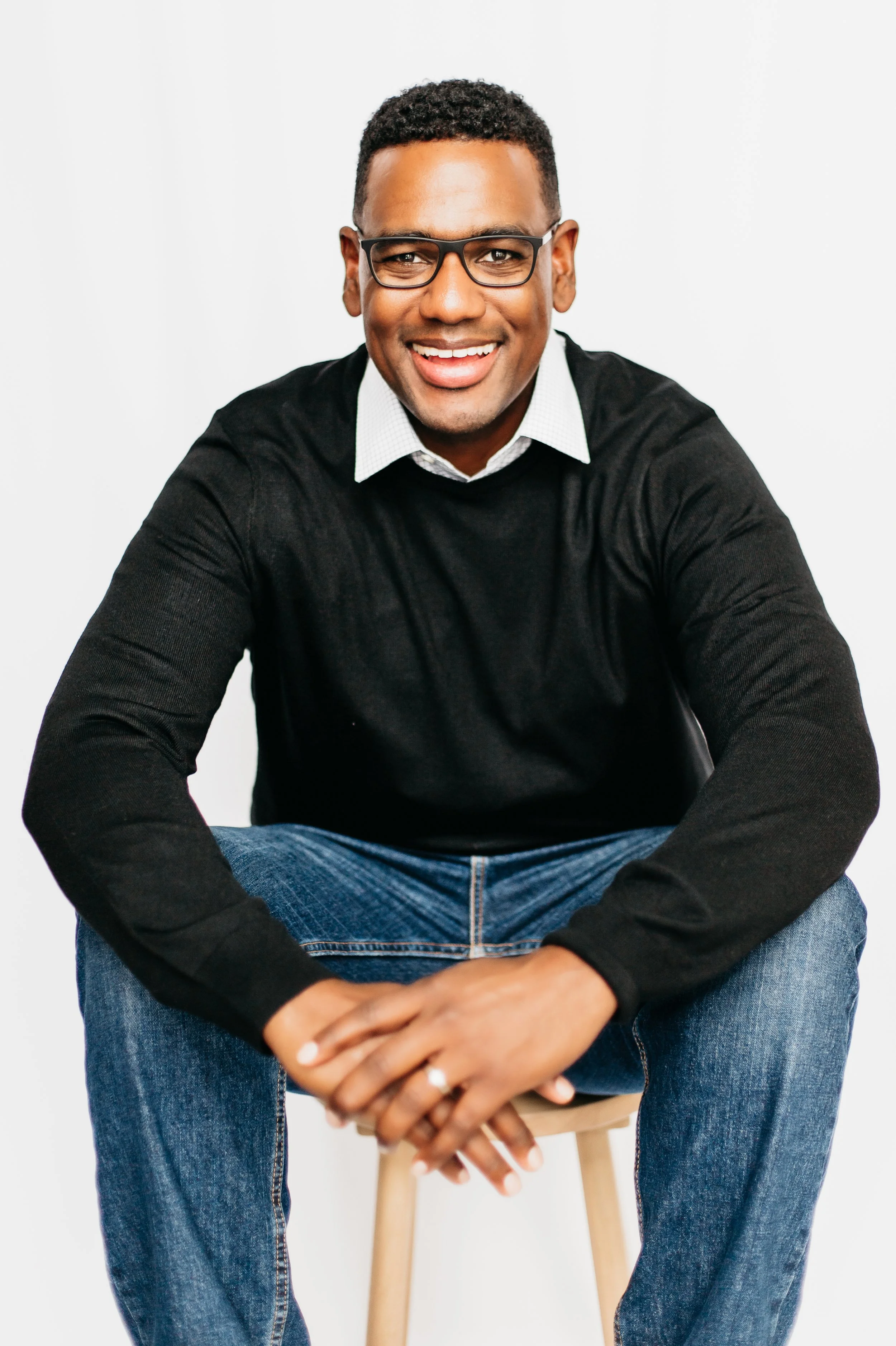 A smiling man wearing glasses, a black sweater, a white collared shirt, and jeans, sitting on a wooden stool against a white background.