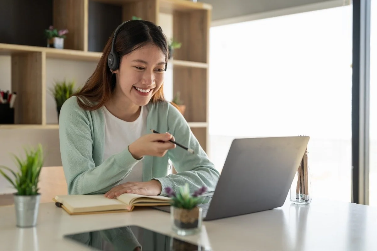 Young woman with headphones smiling, sitting at a desk with a laptop participating in an online Digital Hotpot consultations.