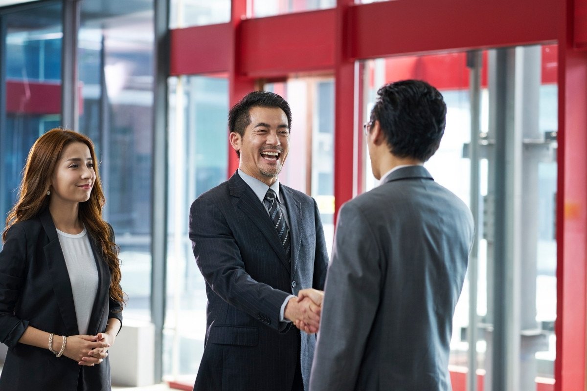 Two businessmen in suits shaking hands and smiling during a professional networking meeting in a modern office, with a female colleague standing nearby.