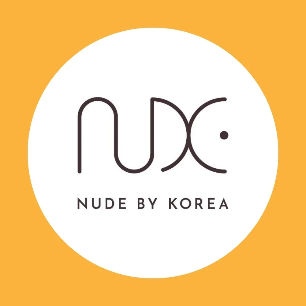 Logo of Nude by Korea, featuring the word 'NUDE' with stylized black lettering and a small dot, on a white circular background with an orange border.