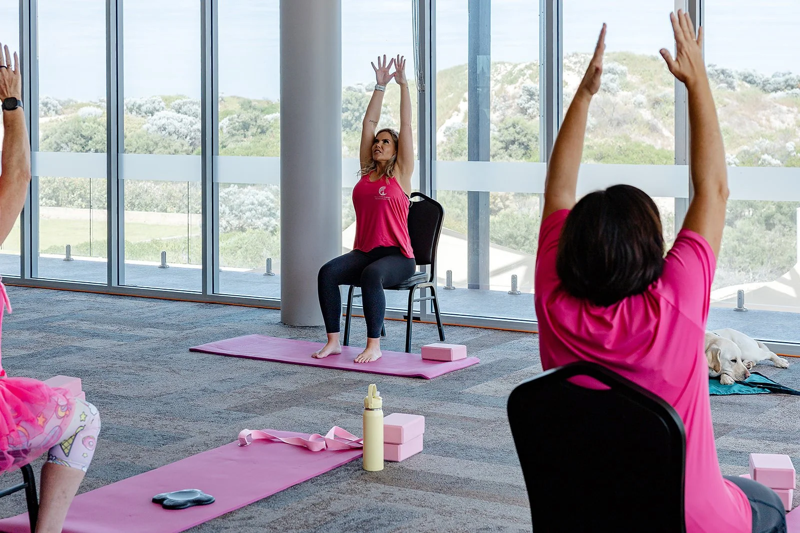 Women participating in a seated yoga or stretching class in a bright studio with large windows showing a scenic outdoor view, with yoga mats, water bottles, and blocks nearby. Yoga with Karney
