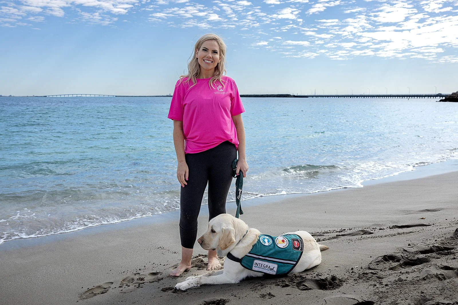 A woman in a pink shirt and black pants standing on a beach with a service dog lying on the sand beside her, ocean and a bridge in the background. Yoga with Karney