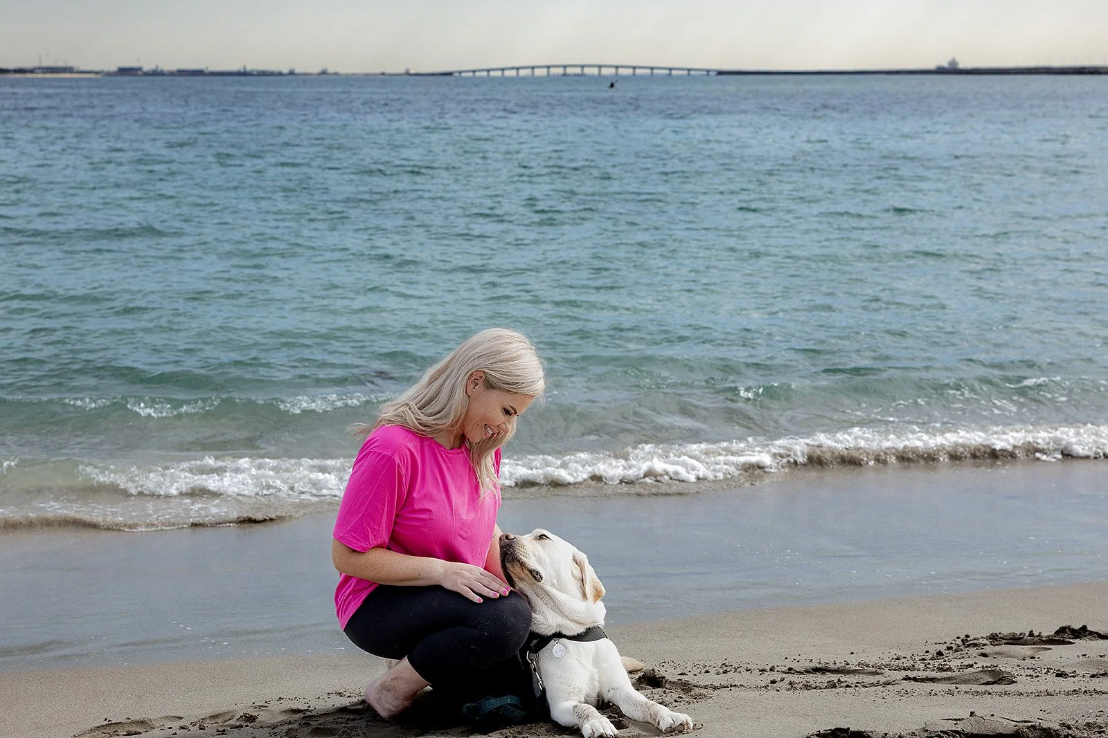 A woman with long gray hair in a pink shirt crouches on the beach, smiling at a Labrador puppy laying in the sand. The ocean with gentle waves and a bridge in the distance are in the background. Yoga with Karney