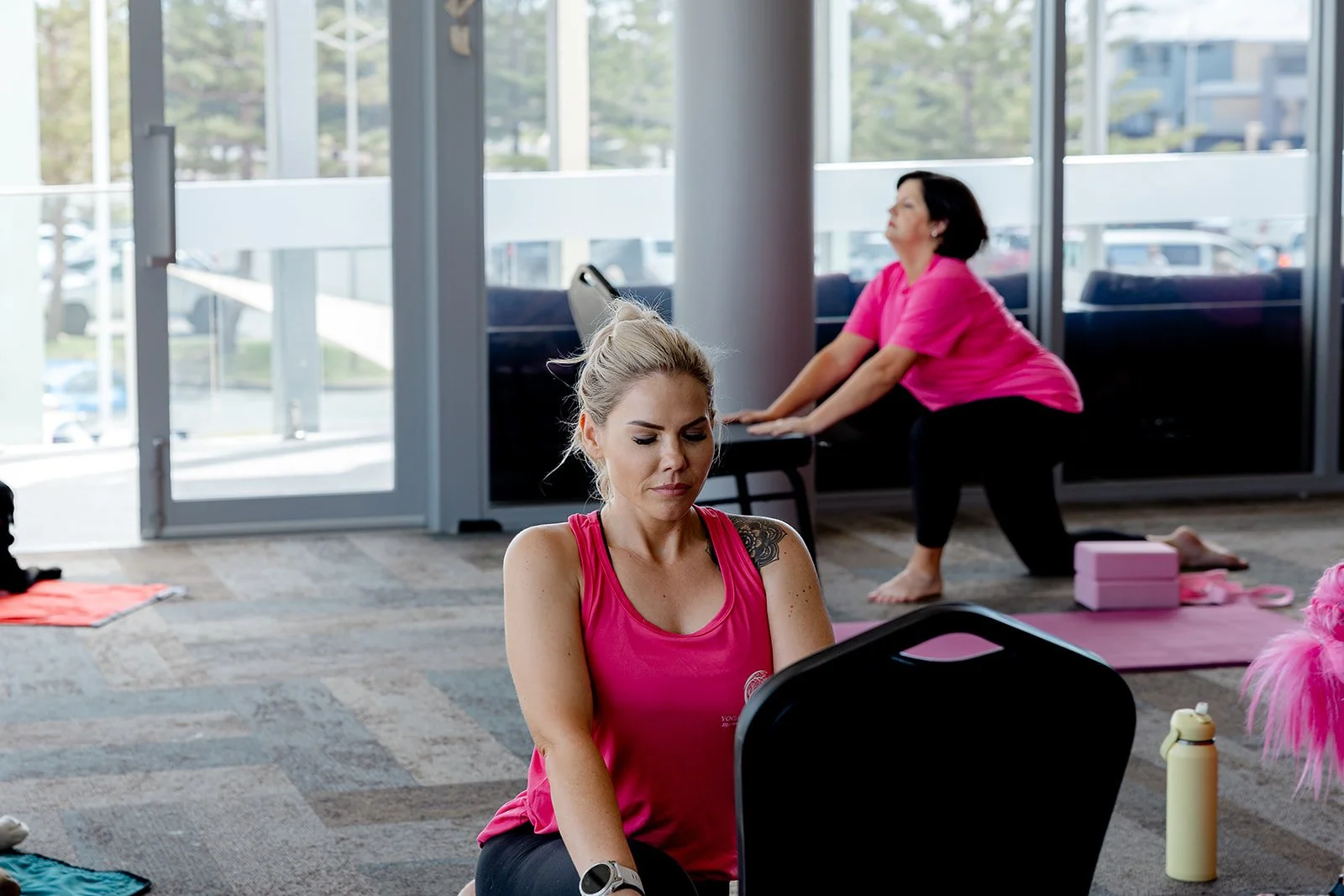 Two women practicing yoga in a studio with large windows. The woman in front is sitting cross-legged and facing forward while the woman in the background is doing a forward bend on a yoga mat. Yoga with Karney