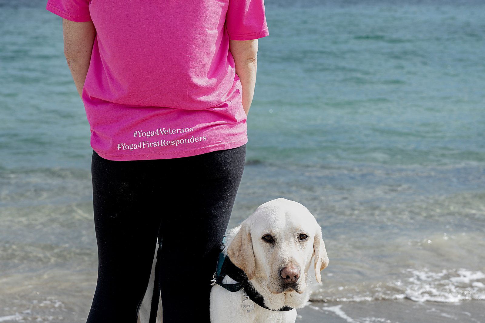 Person wearing a pink shirt with hashtags #Yoga4Veterans and #Yoga4FirstResponders printed on it, standing next to a white Labrador retriever at the beach near the water. Yoga with Karney
