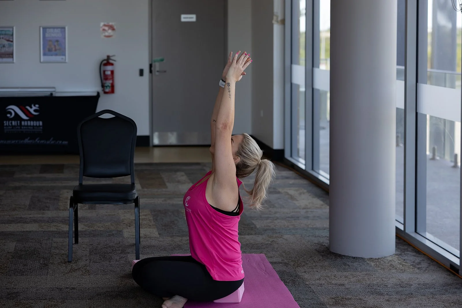 A woman with blonde hair in a ponytail doing a seated yoga stretch on a pink yoga mat in a room with large windows, a black chair, and a fire extinguisher in the background. Yoga with Karney