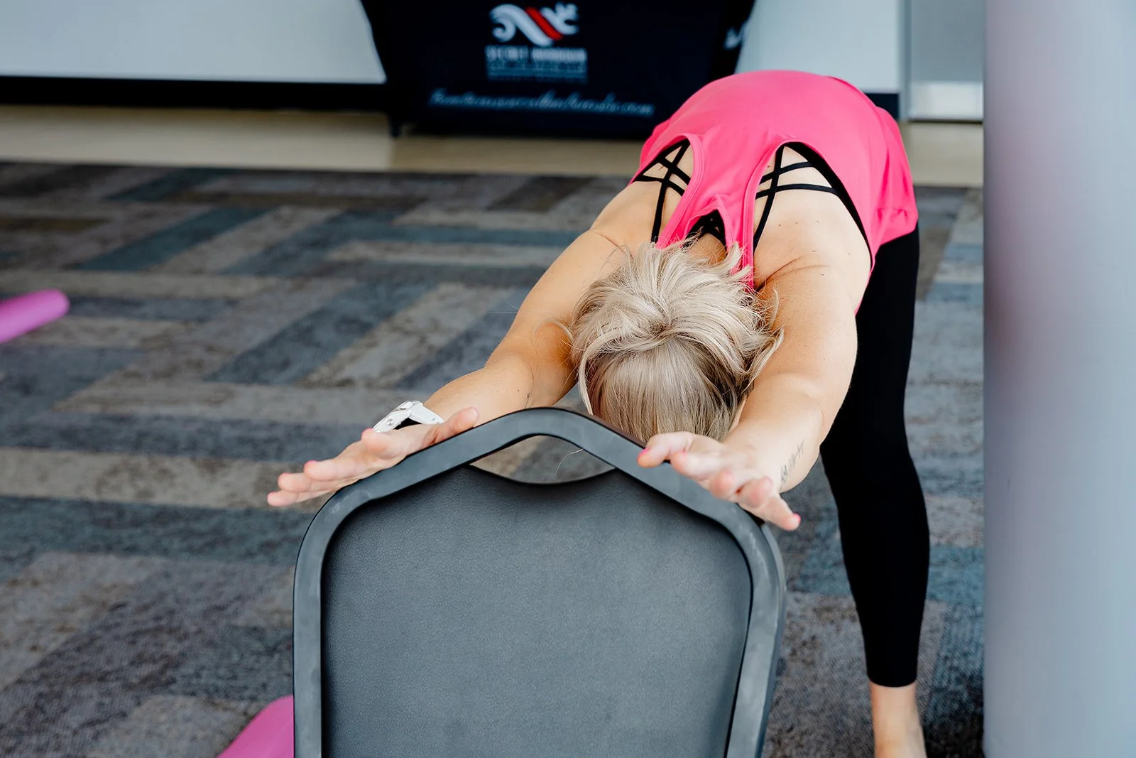 A woman in athletic clothing doing a child's pose stretch on a chair in a fitness or yoga studio. Yoga with Karney