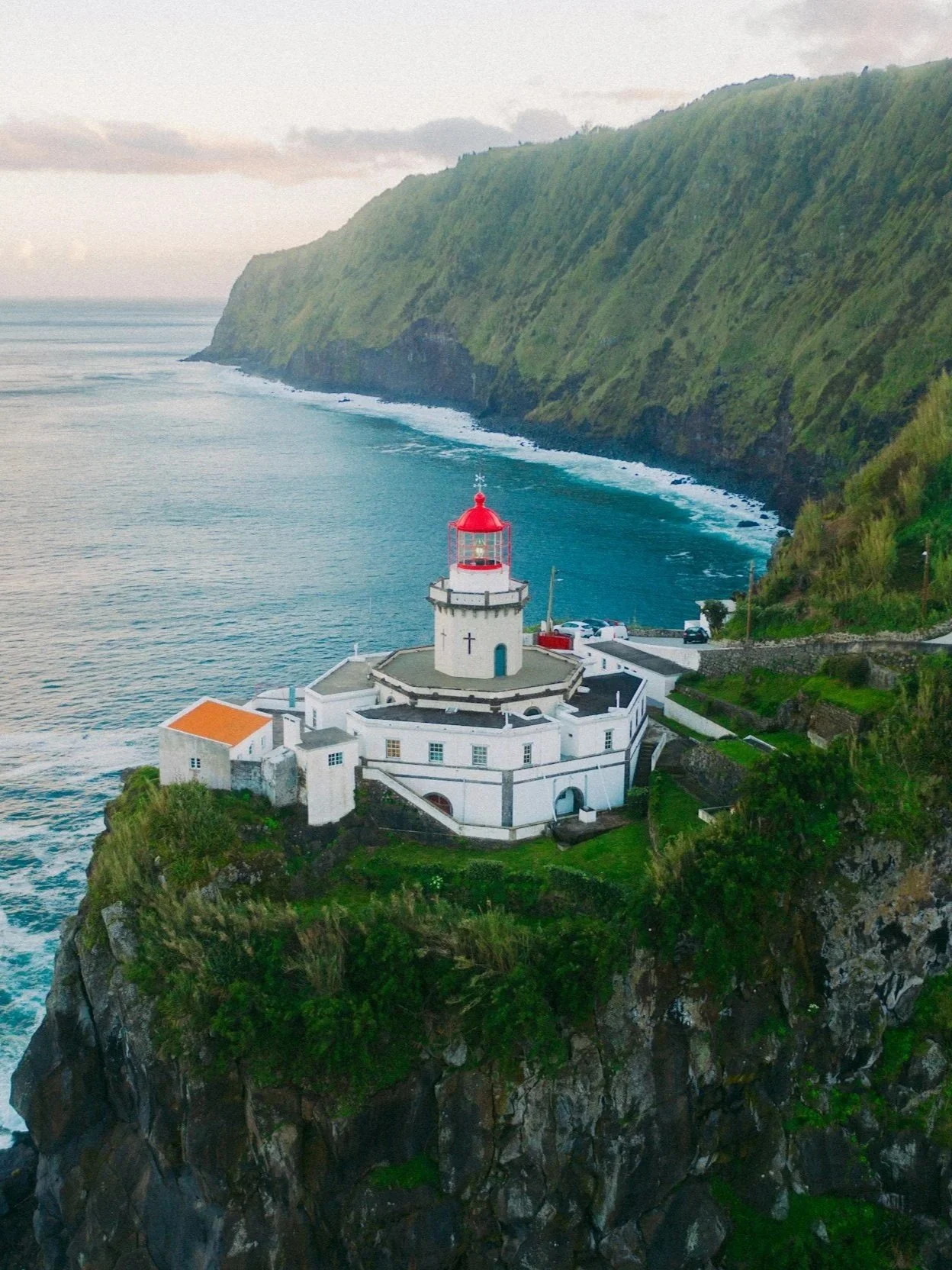 Leuchtturm auf einer Klippe mit grünem Berg im Hintergrund und dem Meer im Vordergrund.