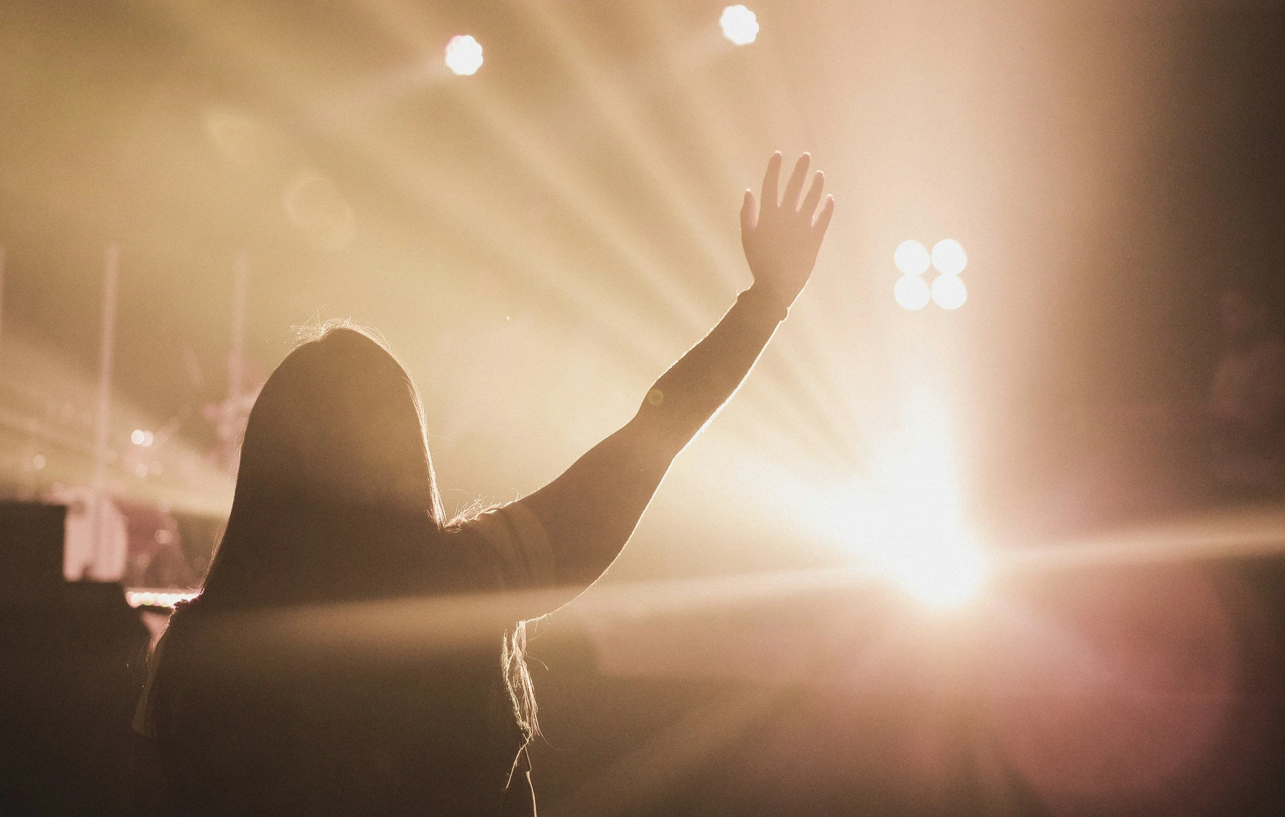 A person raising their hand in front of bright stage lights, possibly at a concert or event.