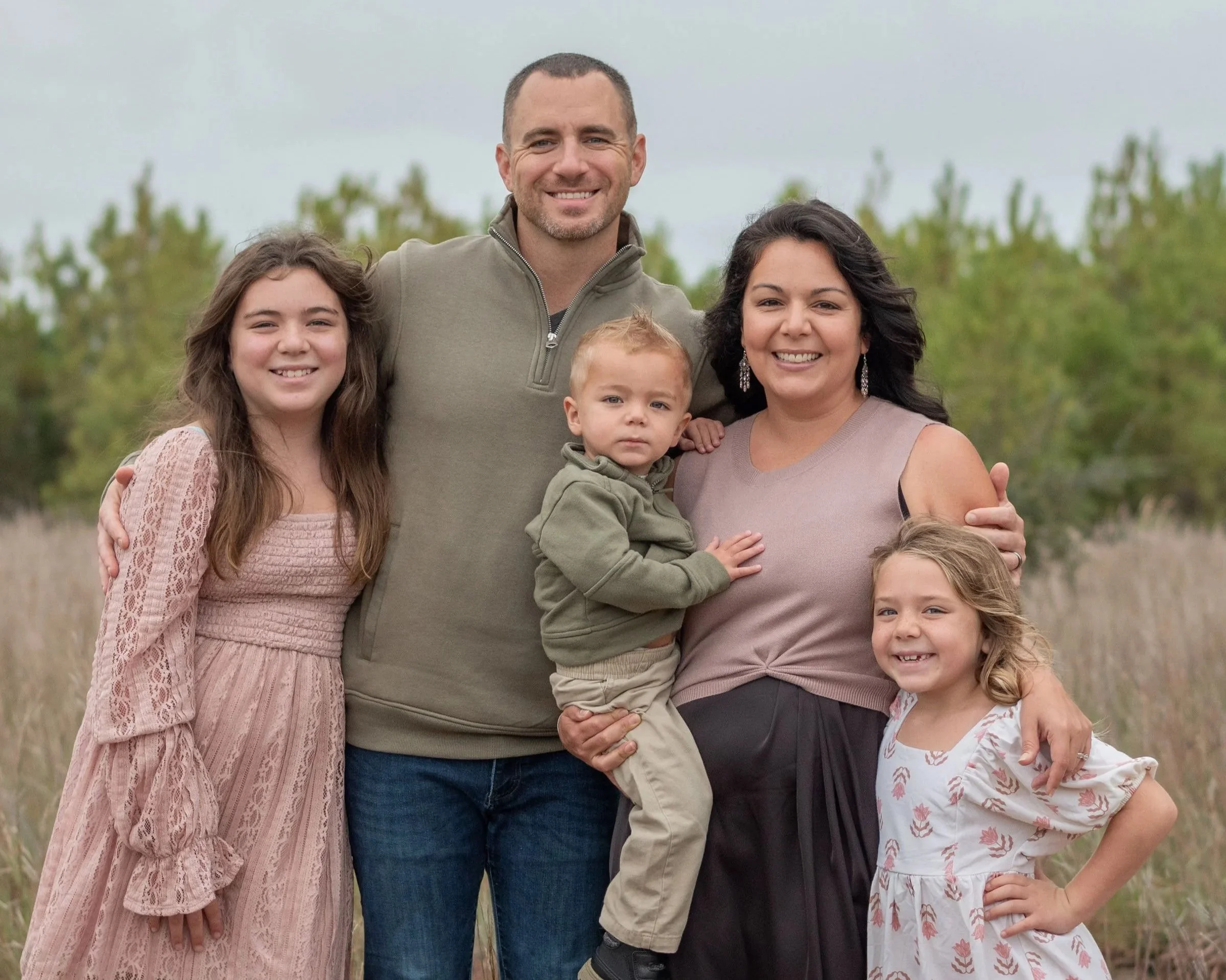 Outdoor family portrait of TJ Bowser and his family, standing on grass with trees and greenery in the background. Taken for Gospel Fellowship Church in Utah, representing faith, family, and community leadership.